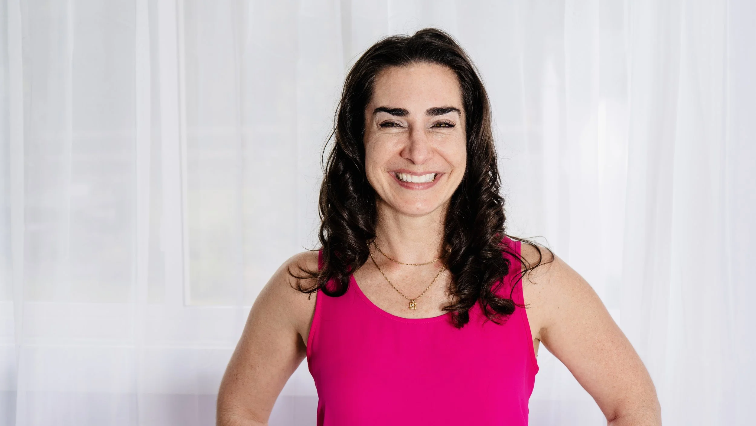 A woman with dark, curly hair smiling, wearing a bright pink sleeveless top and layered gold necklaces, standing in front of a white curtain background.