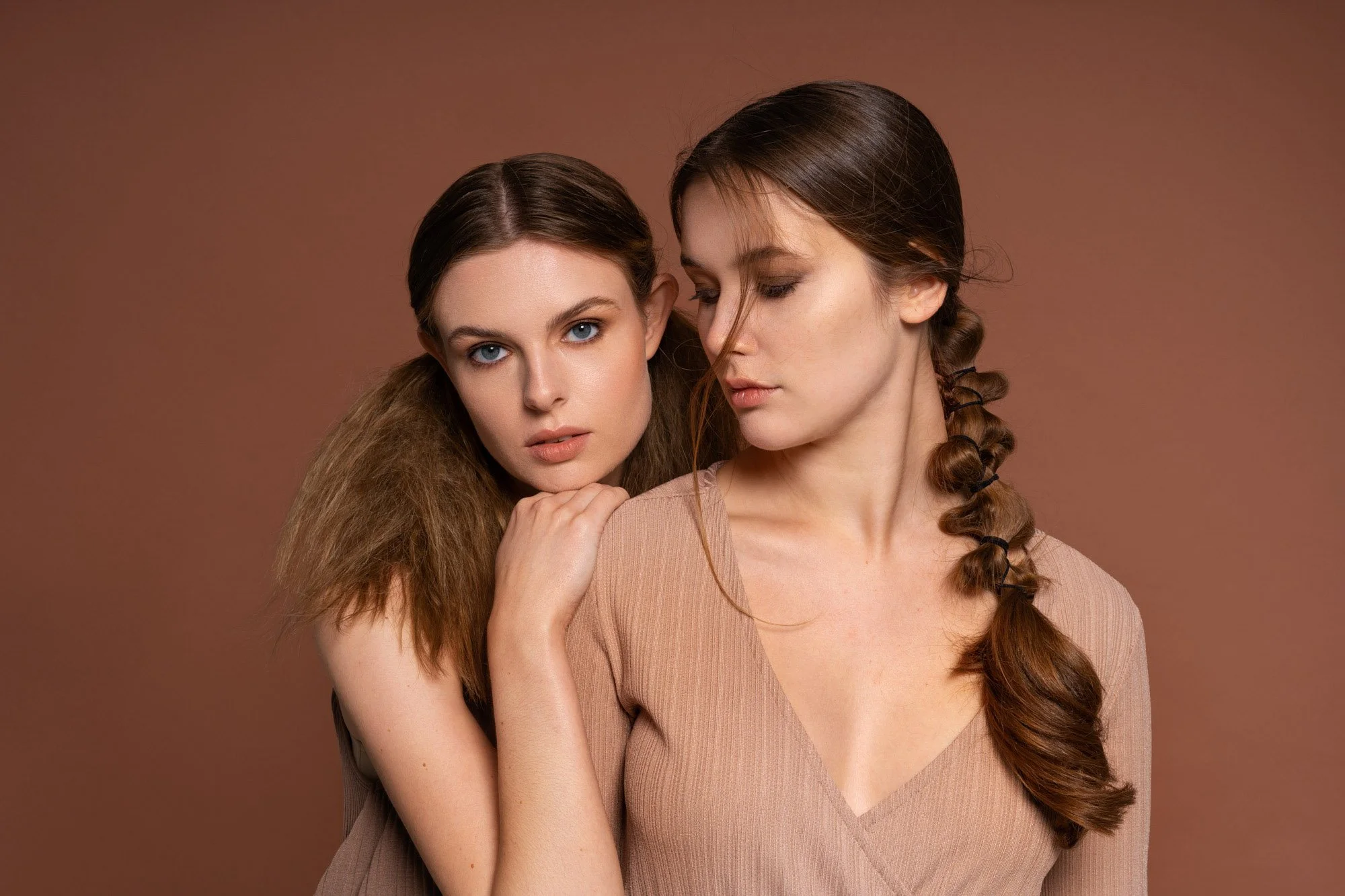 Two women with brown hair and fair skin posing against a plain brown background, one with her hand on the other's shoulder.