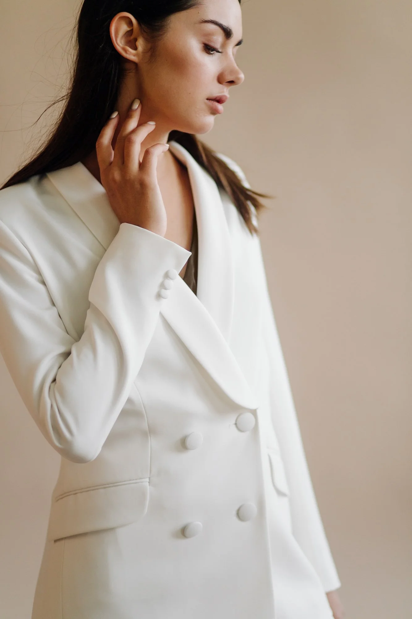 A woman with dark hair dressed in a white blazer with matching pants, looking to the side with her hand touching her neck.