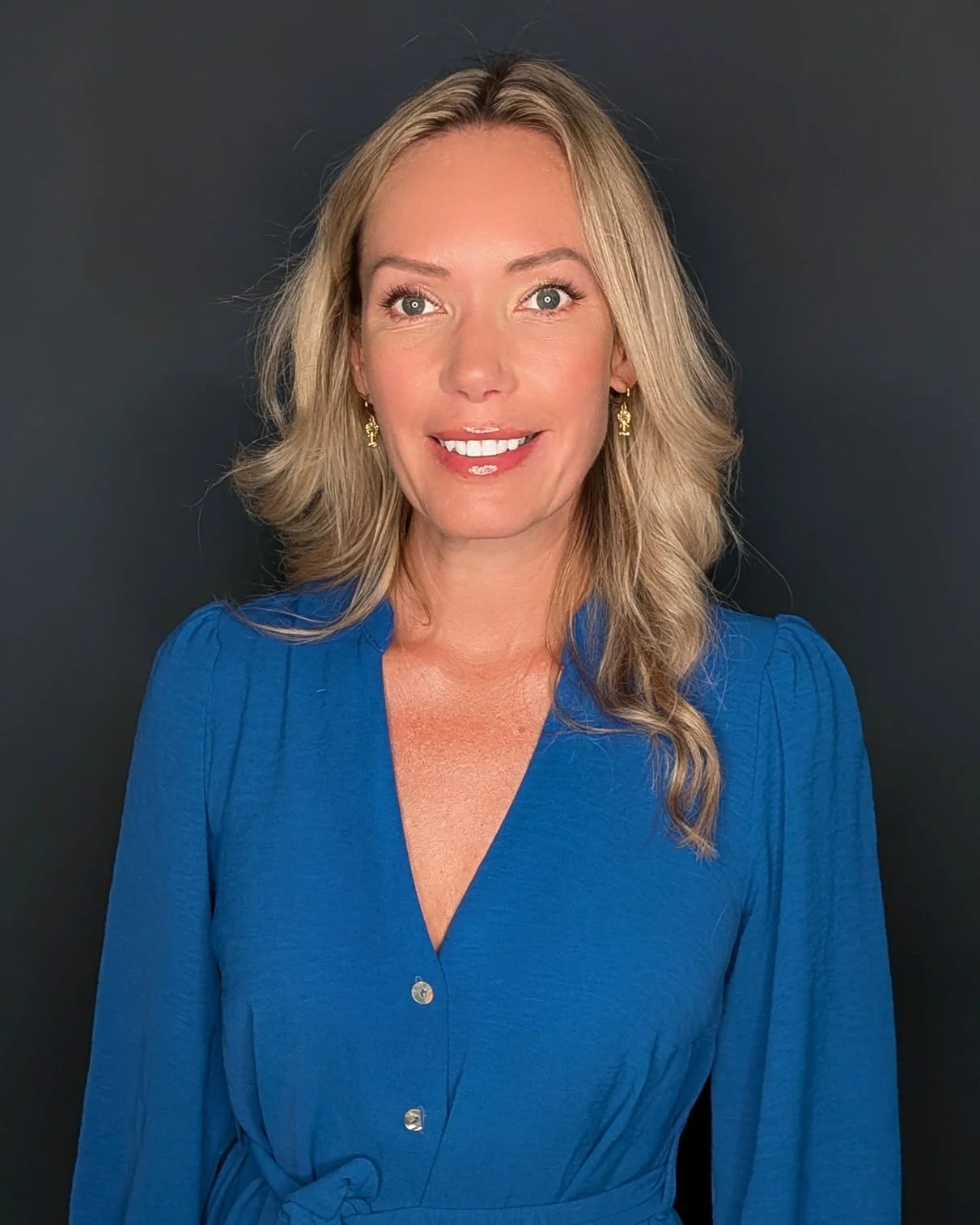 A woman with blonde wavy hair, blue eyes, and fair skin smiles in front of a dark background. She is wearing a bright blue dress with buttons and gold earrings.