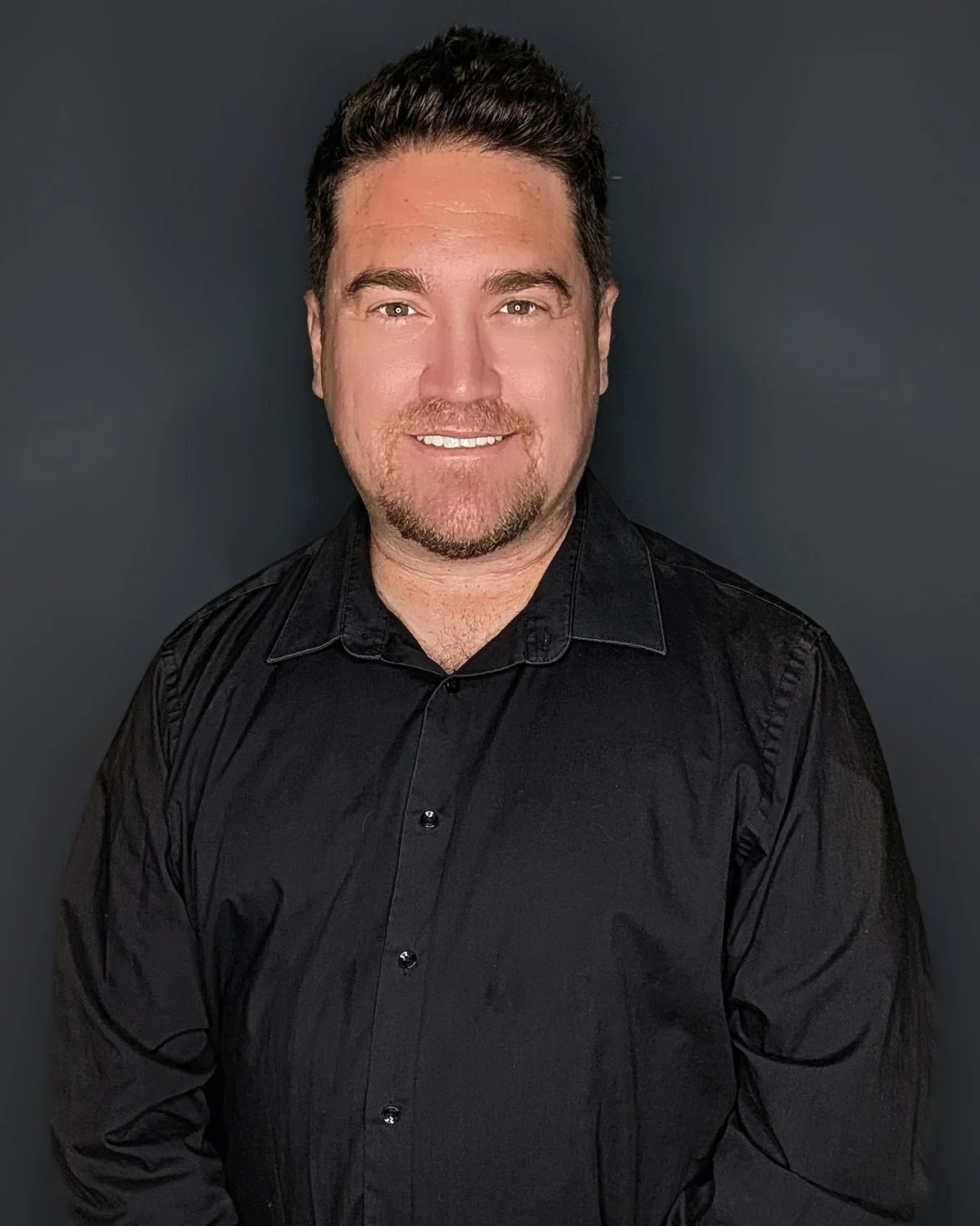 Headshot of a man with short dark hair, light skin, and a goatee, wearing a black collared shirt, smiling, against a dark gray background.