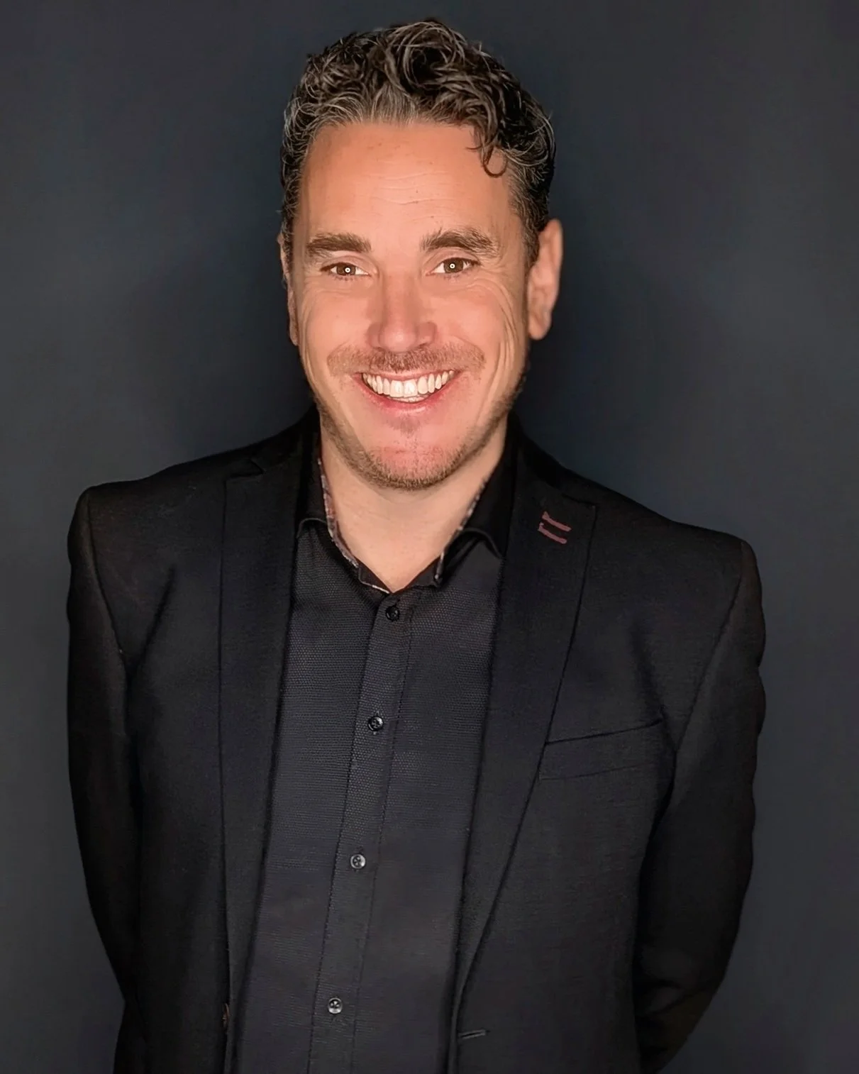 A smiling man with curly hair and a beard, wearing a black suit and black shirt, standing against a dark background.