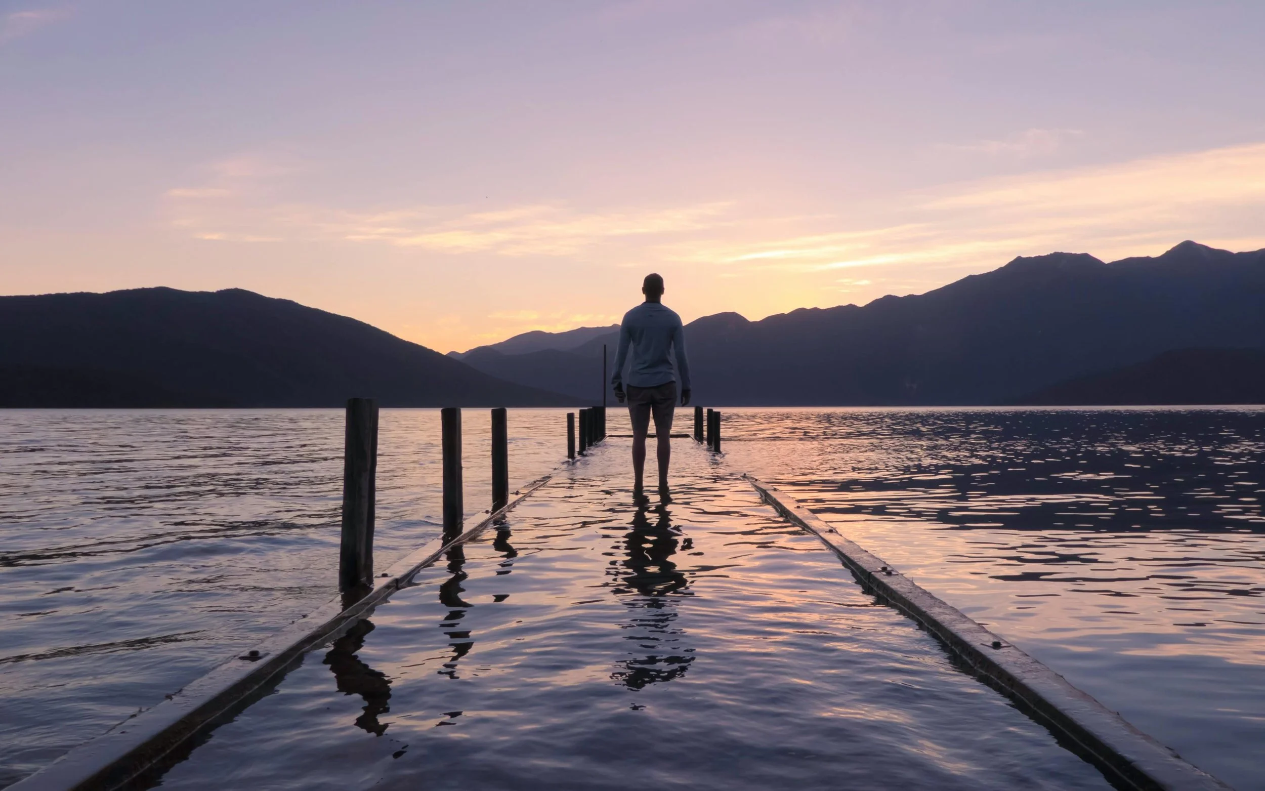 A solitary person walking along a flooded pier at sunset—symbolising the quiet inner struggle of self-doubt and the path toward greater self-acceptance and clarity.