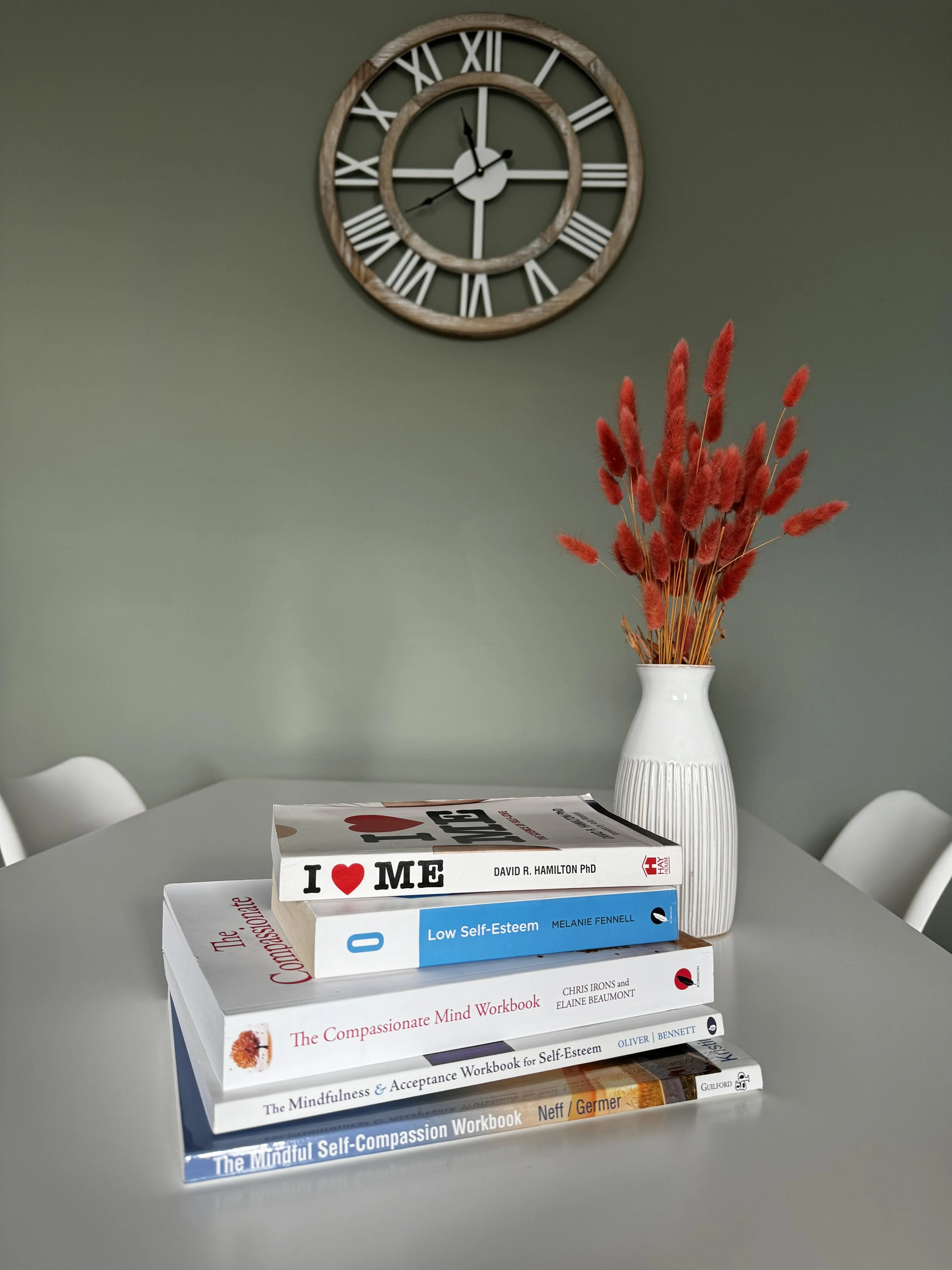 A curated stack of self-help and self-compassion books beside a vase of red florals—symbolising growth, healing, and the journey to realignment and flourishing.