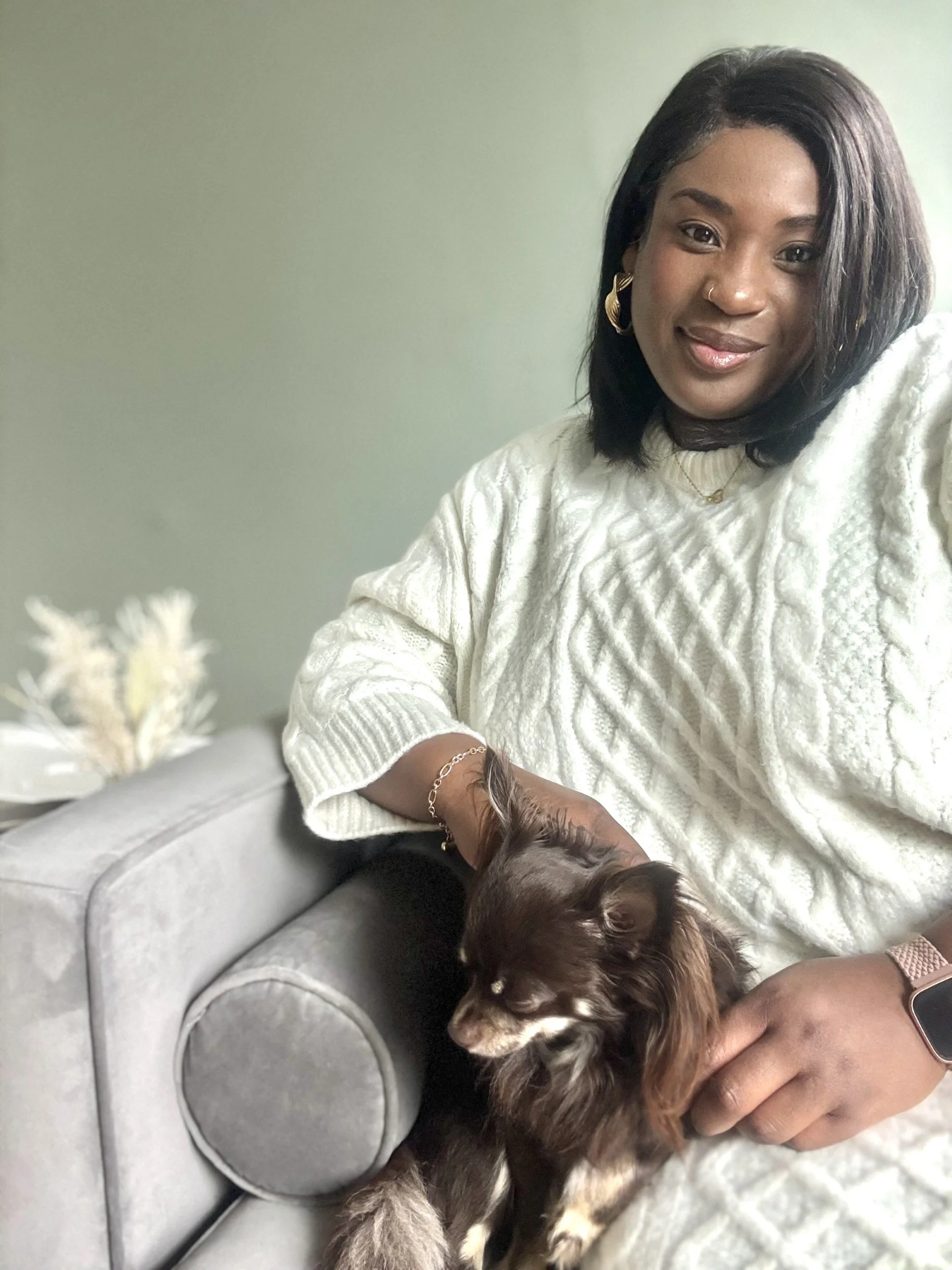 A woman with dark hair and gold jewelry sitting on a gray couch, holding a small brown dog with white markings.