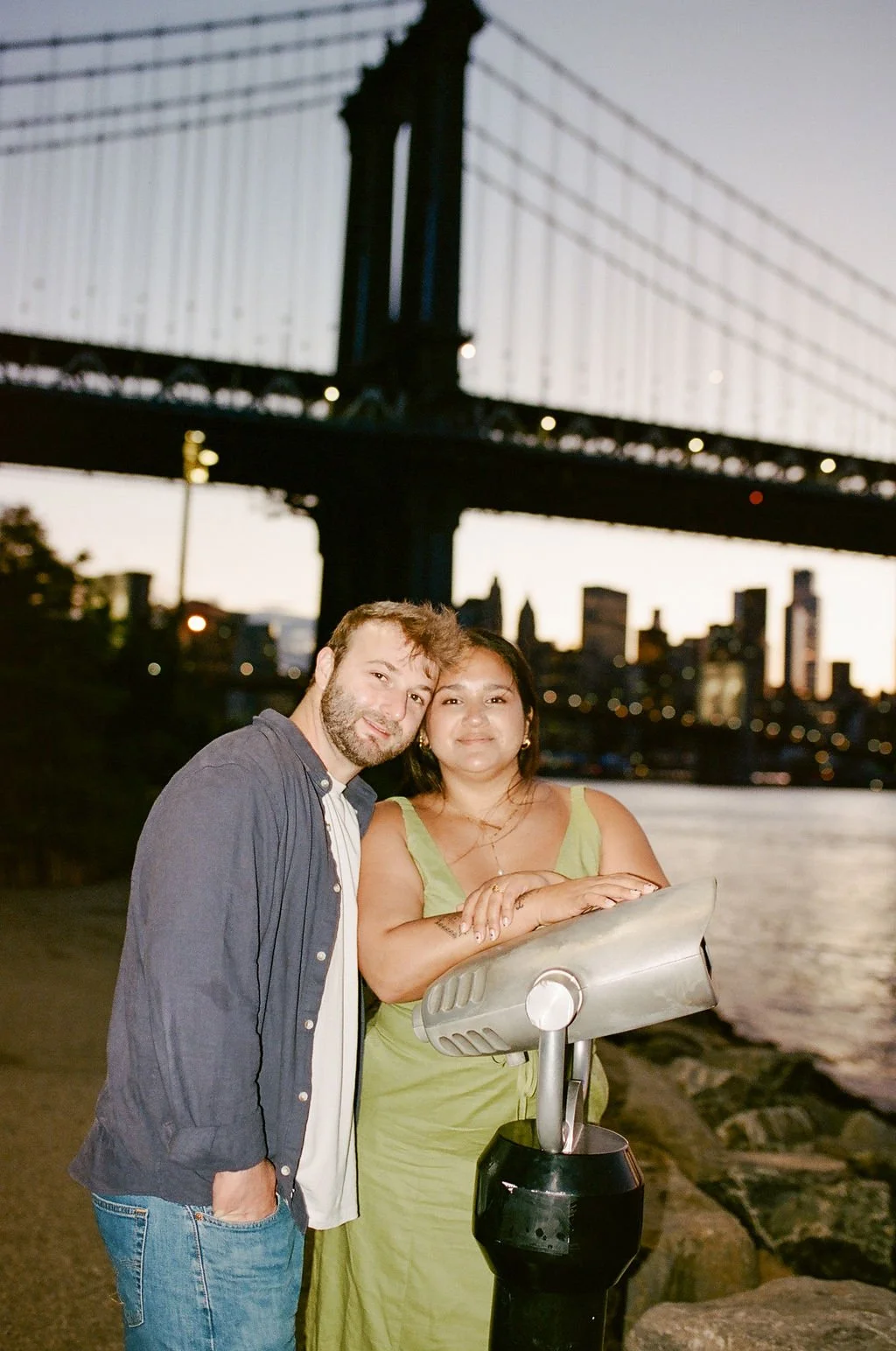 A man and woman standing next to each other by New York City harbor, with the Brooklyn Bridge and city skyline in the background during sunset.