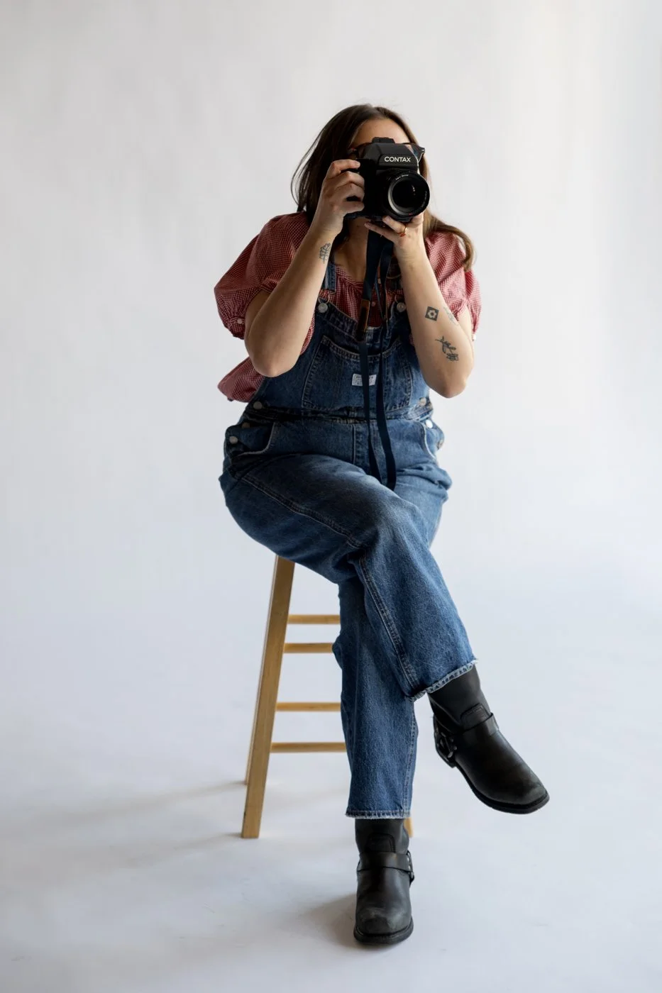 Delaney Hady sitting on a wooden stool, holding a camera up to her face, wearing denim overalls, a red checked shirt, and black boots, with tattoos on her arms, against a plain white background.