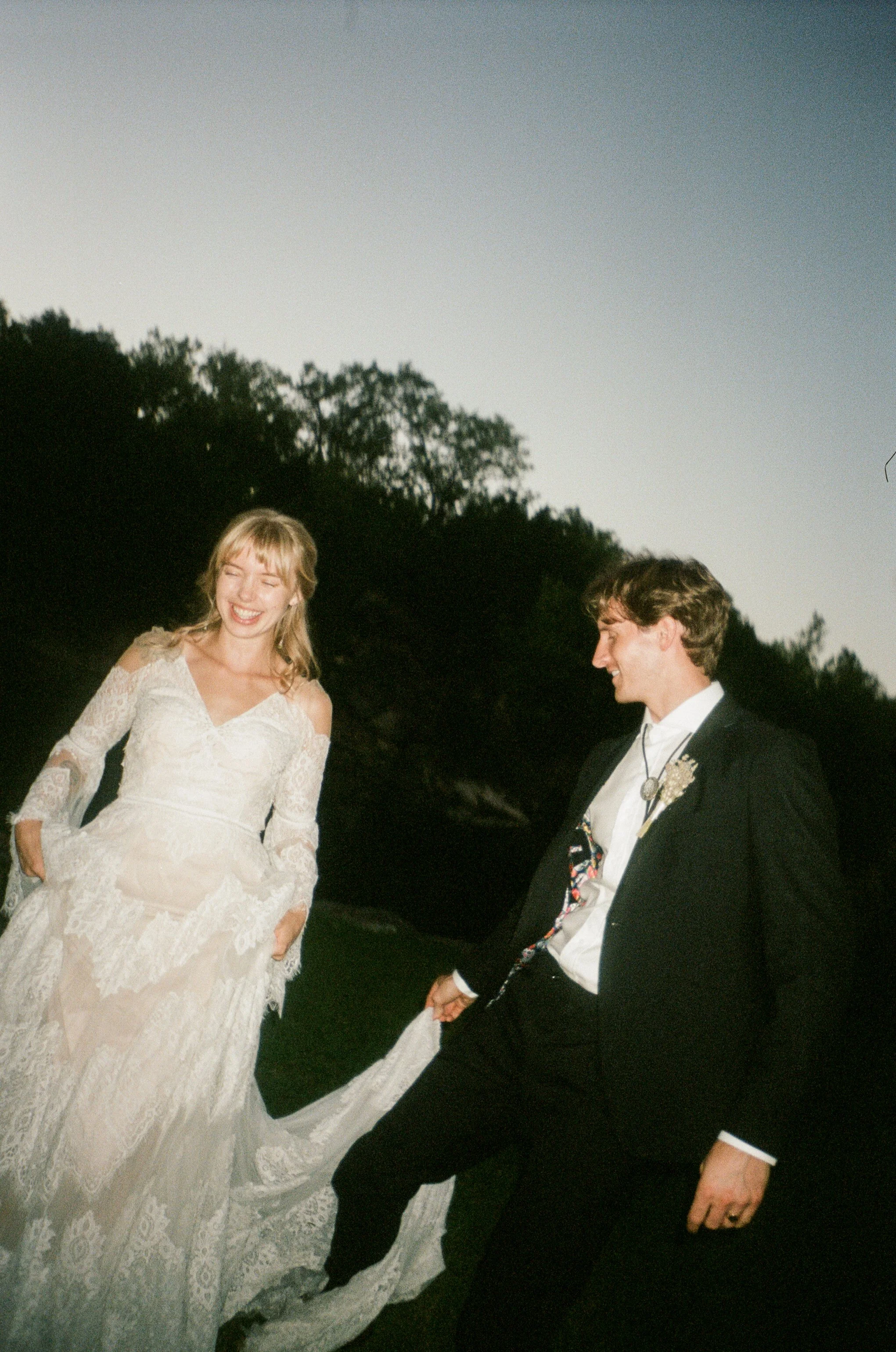 A film photo of a smiling bride in a lace wedding dress holding hands with a groom in a black suit and tie outdoors during dusk or evening.