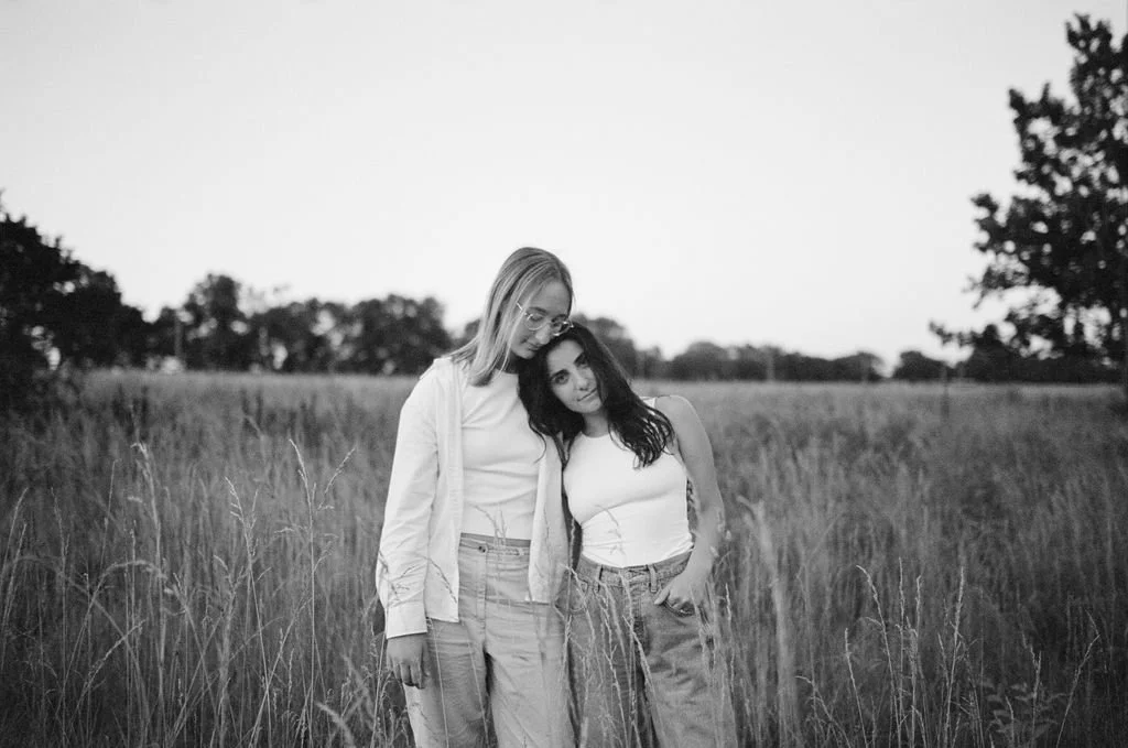 A queer couple standing in tall grass in a field with trees in the background, one resting her head on the other's shoulder, both looking at the camera, in black and white.