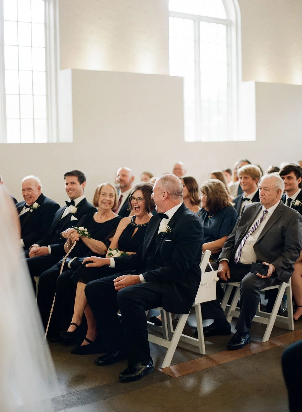 Guests at a wedding seated in a bright, spacious hall with high windows, smiling and enjoying the ceremony.