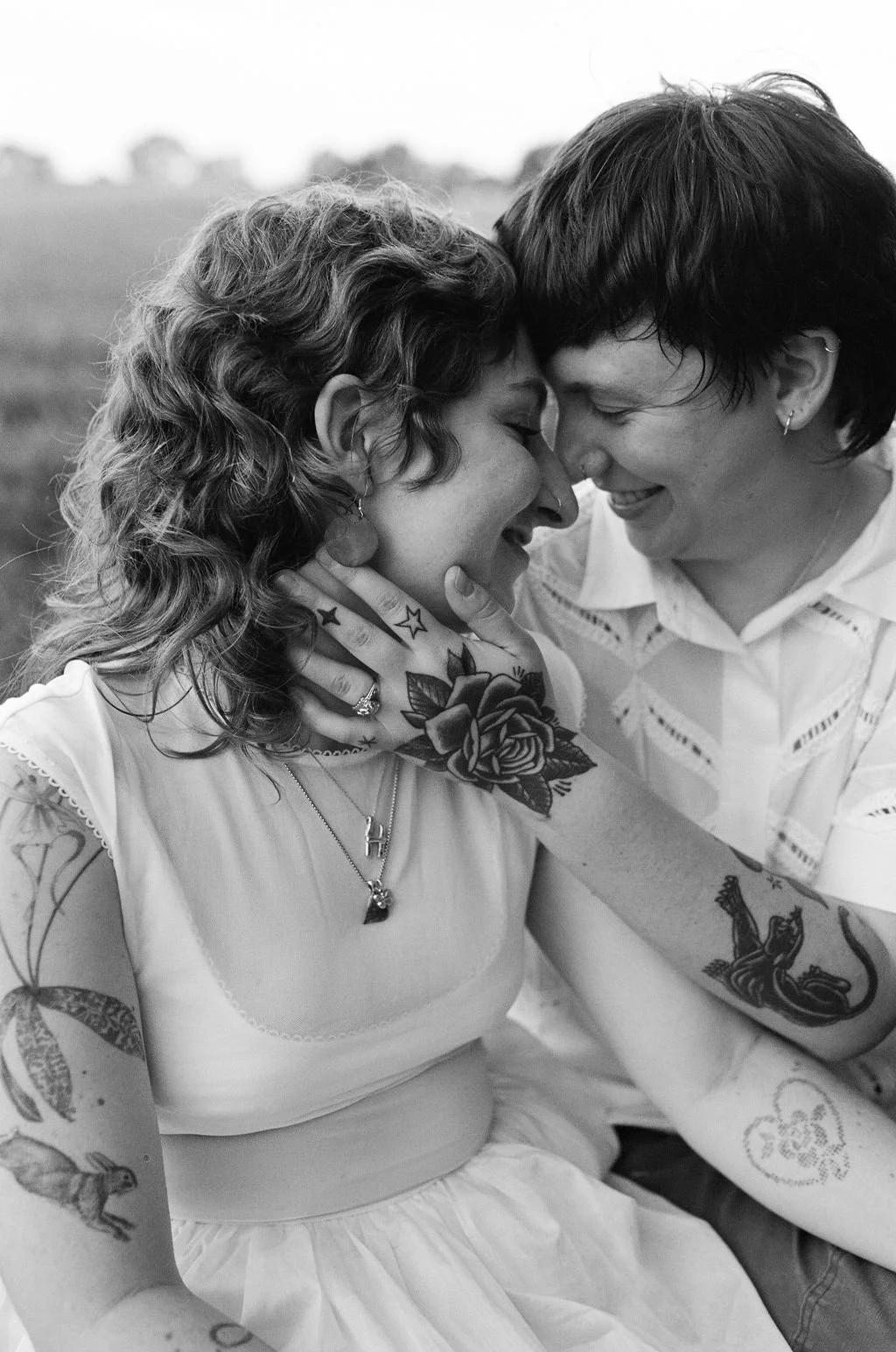 A queer couple with tattoos touching foreheads and smiling at each other in an outdoor setting.