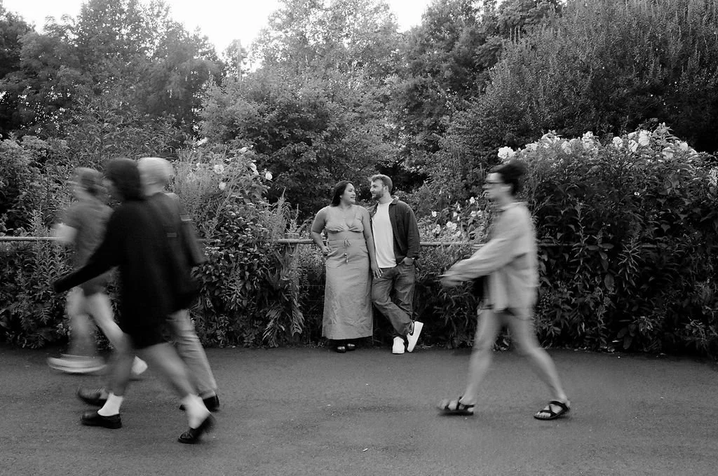 A black and white photo of five people in a park, with two standing against a bush background and three walking on the path in the foreground.
