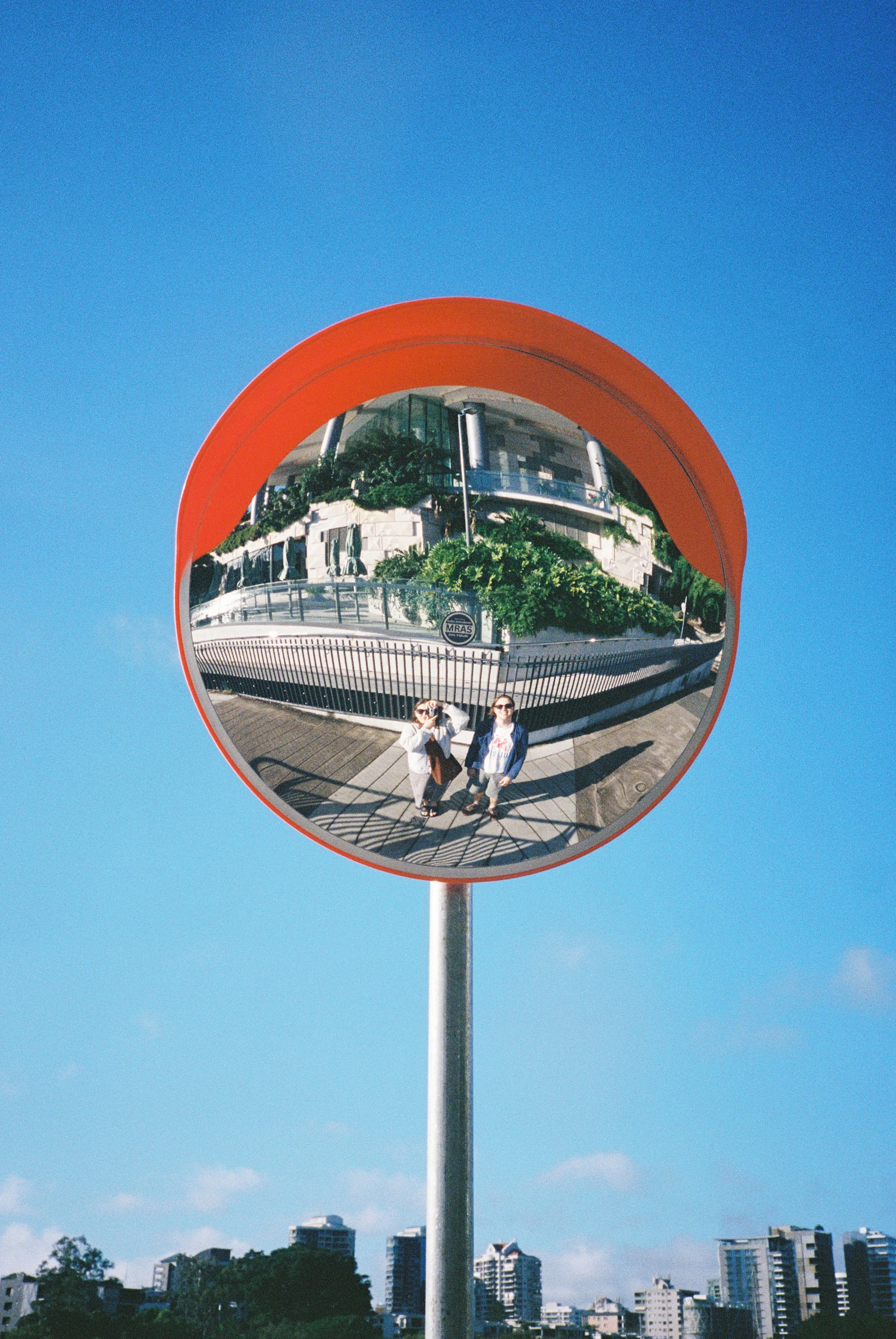 A circular mirror on a pole reflecting two people taking a photo, with a building, greenery, and cityscape in the background against a blue sky.