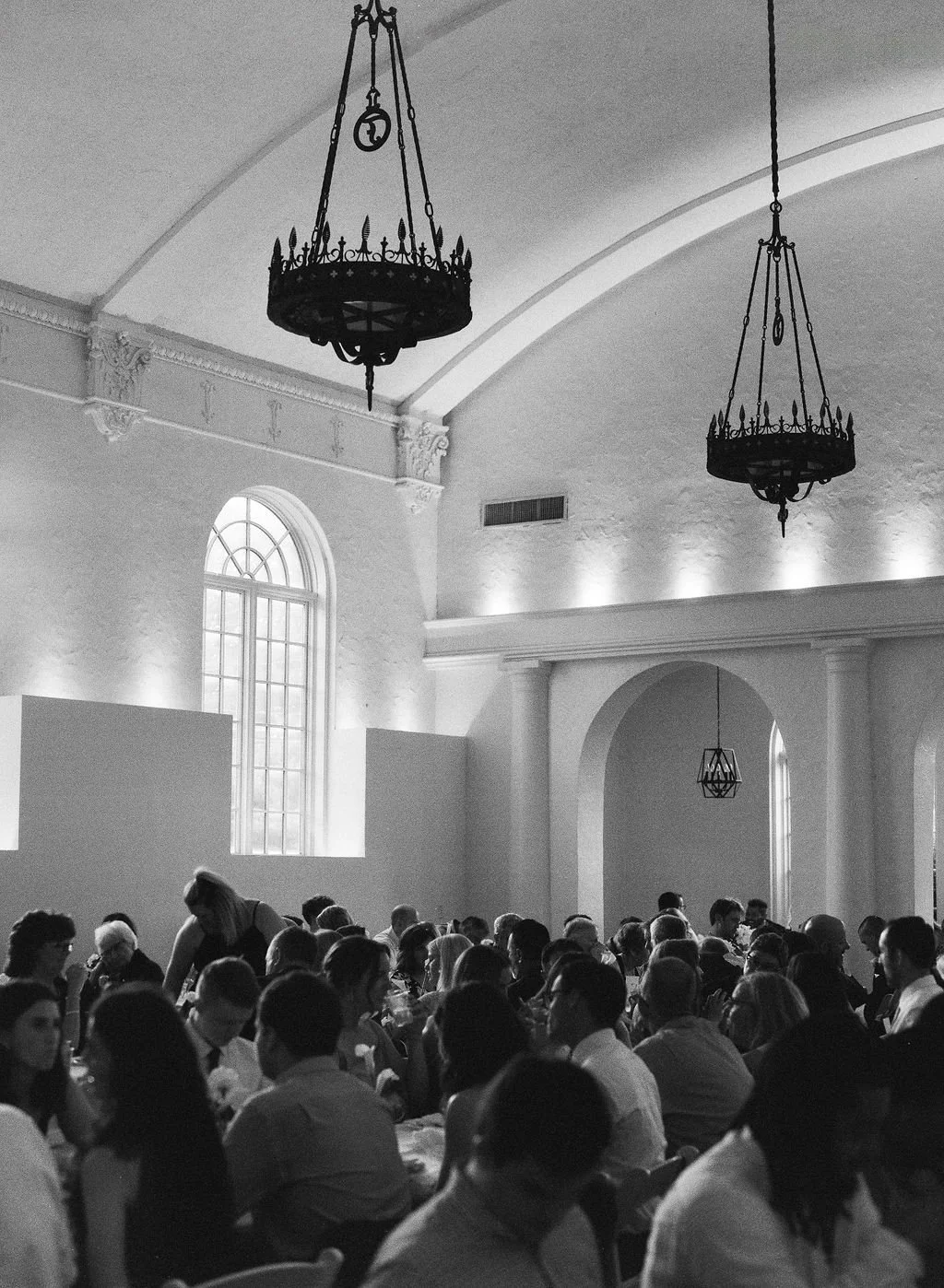 A black and white photo of a large gathering of people seated at tables inside a spacious room with high ceilings, arched windows, and decorative chandeliers.