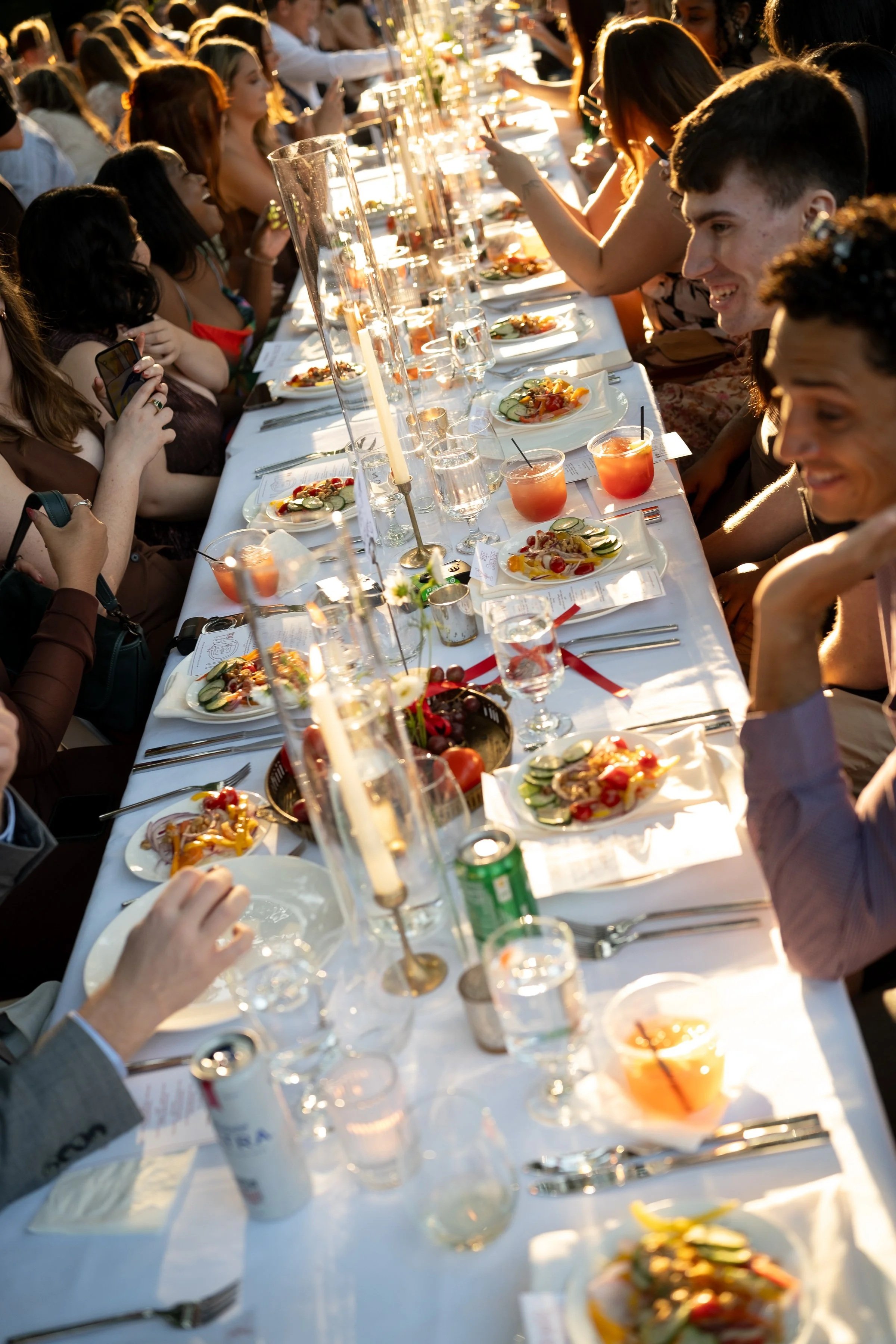 A long banquet table set for a celebration with many guests dining, drinking, and taking photos in sunlight.