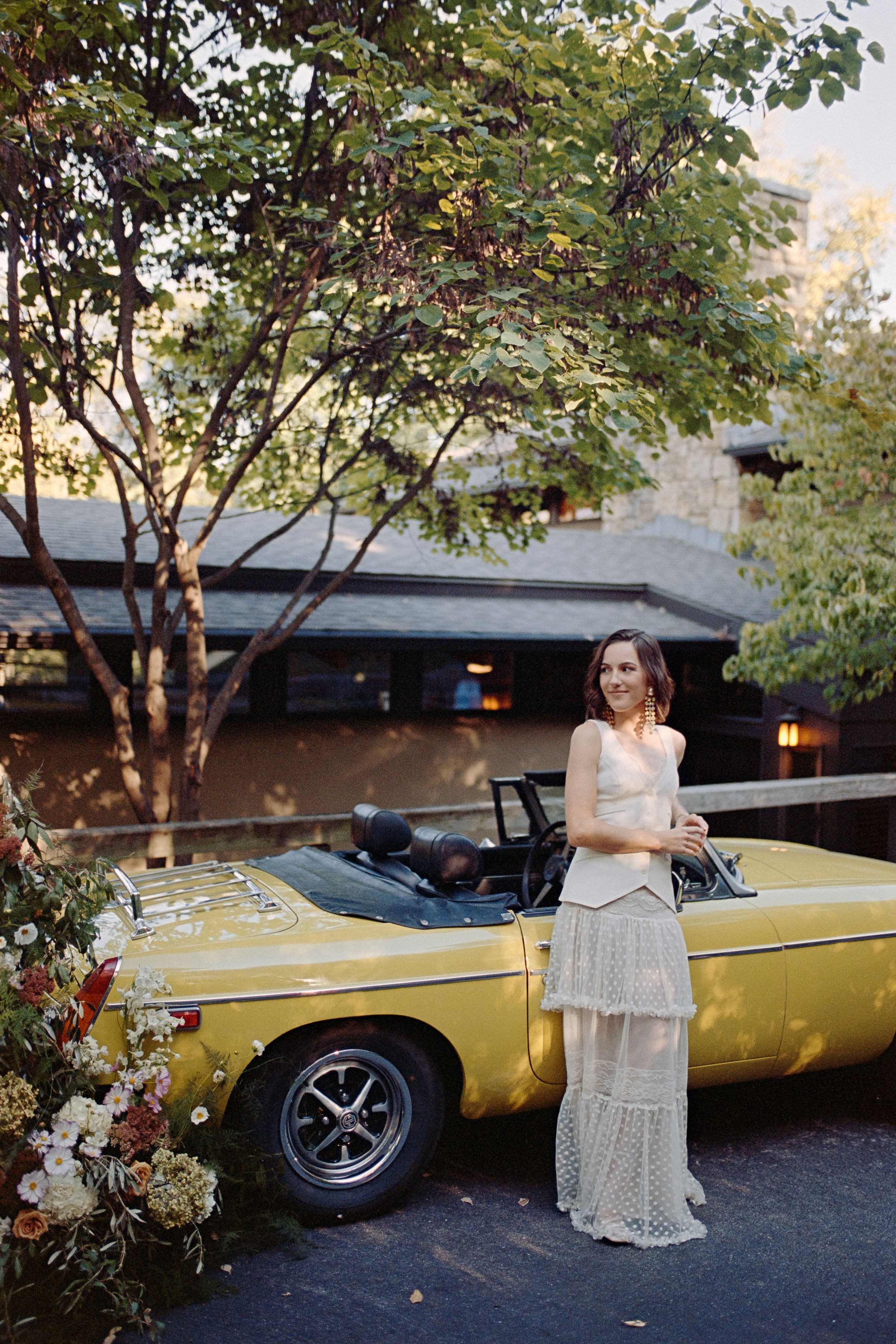 Film photograph of a bride in her fashion forward wedding attire. She is standing in front of a vintage yellow car with a floral arrangement.