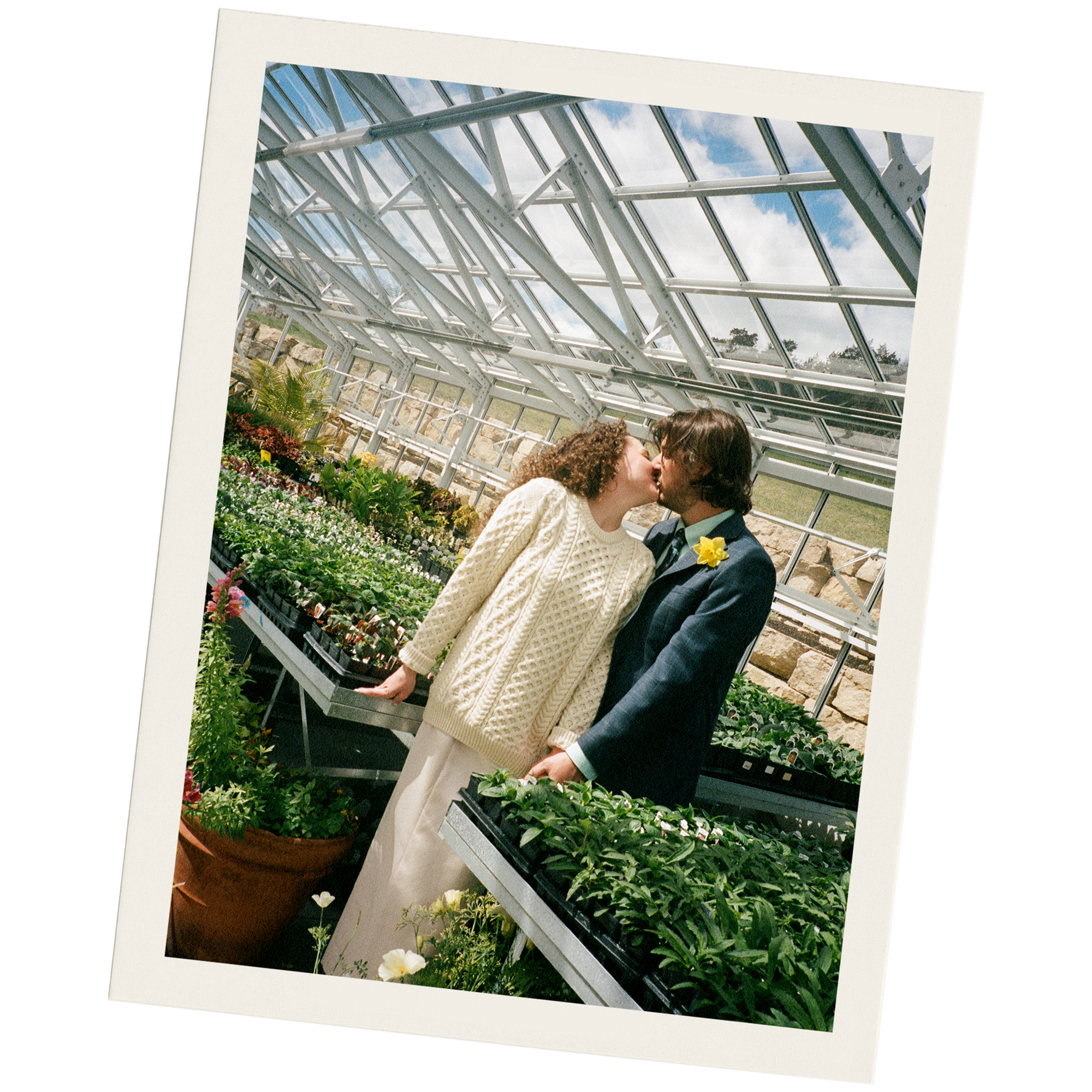 A couple kissing inside a greenhouse with potted plants and flowers around them.