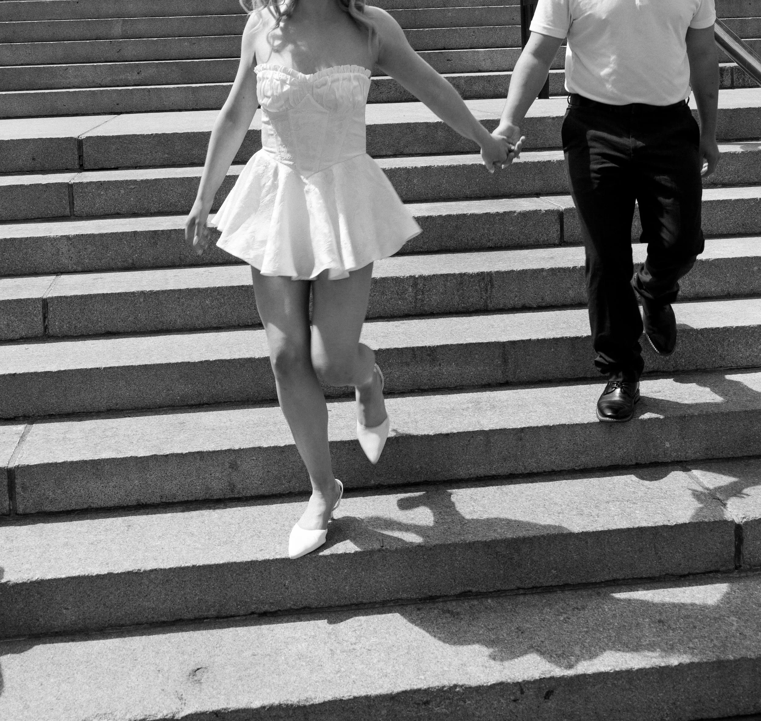 A woman in a short, ruffled dress and high heels holding hands with a man in dark pants and a light shirt, walking down outdoor stairs.