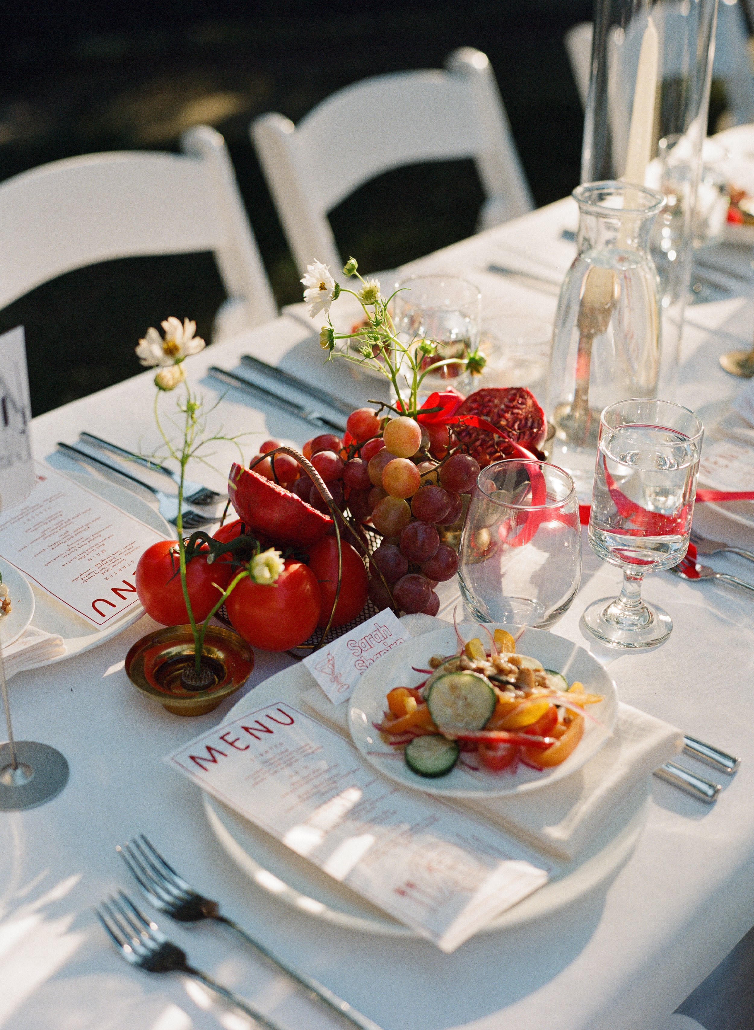 A table set for a meal with a centerpiece of grapes, tomatoes, and flowers, along with plates of salad, menus, and glassware, on a white tablecloth with white chairs.