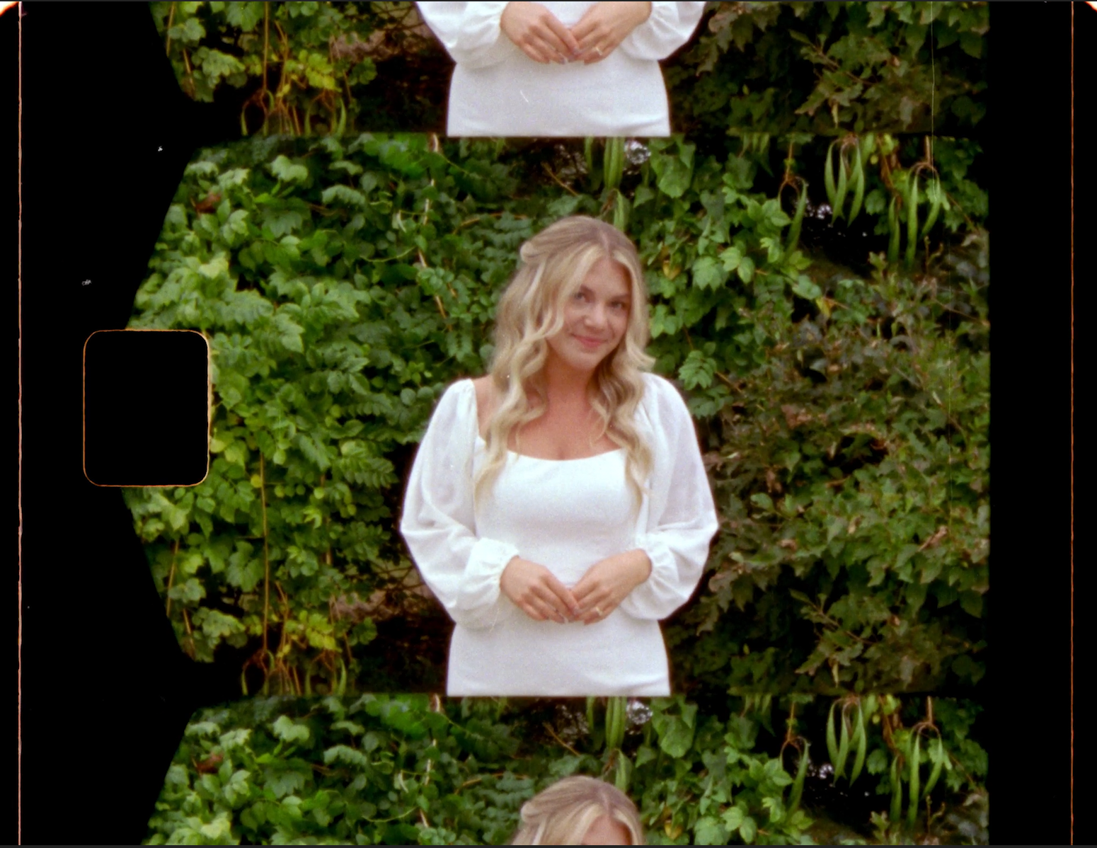 A woman with long blonde hair in a white dress outdoors, with green bushes in the background.