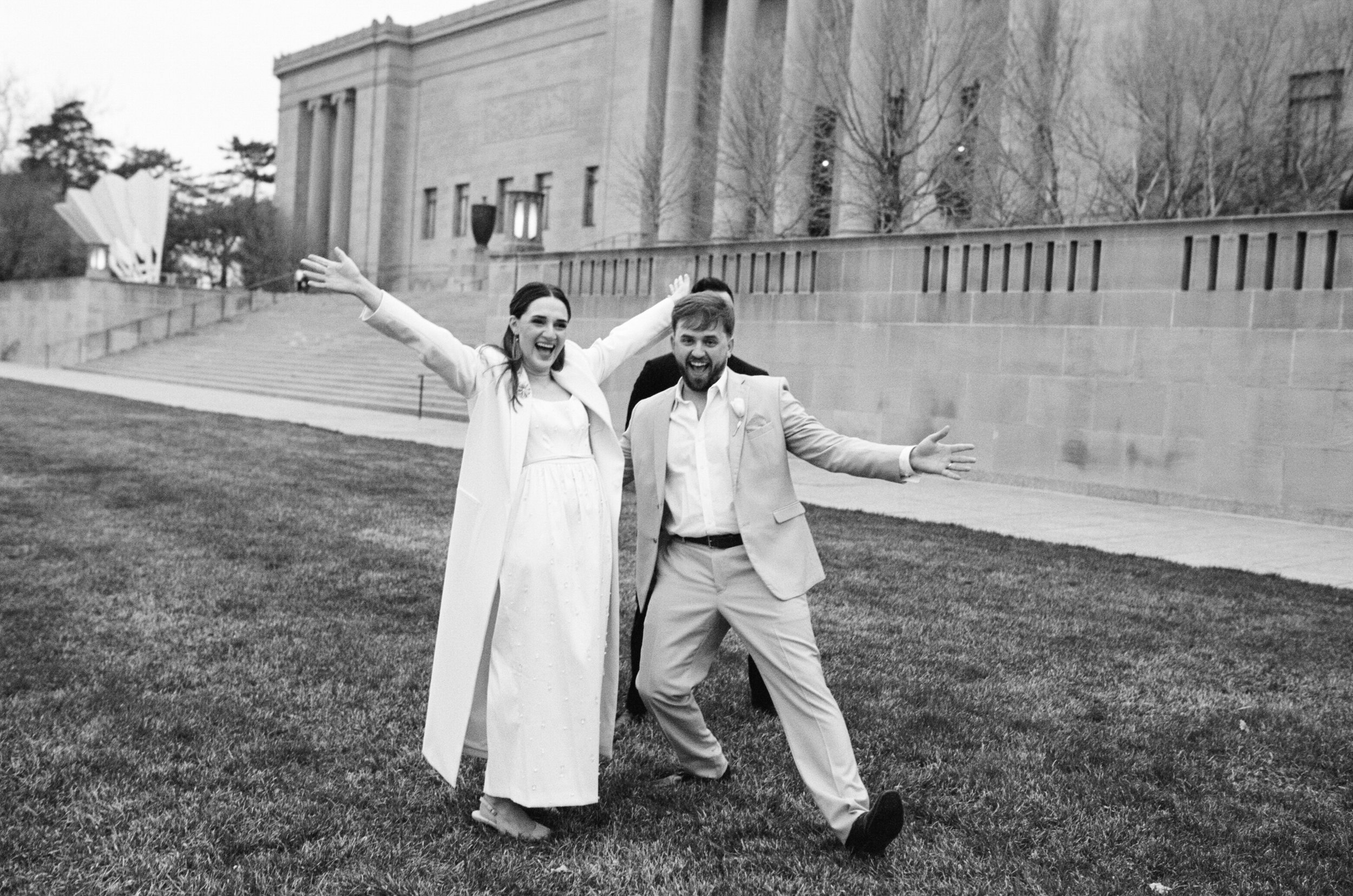 A couple celebrating getting married with their arms stretched wide in front of the Nelson Atkin Museum.