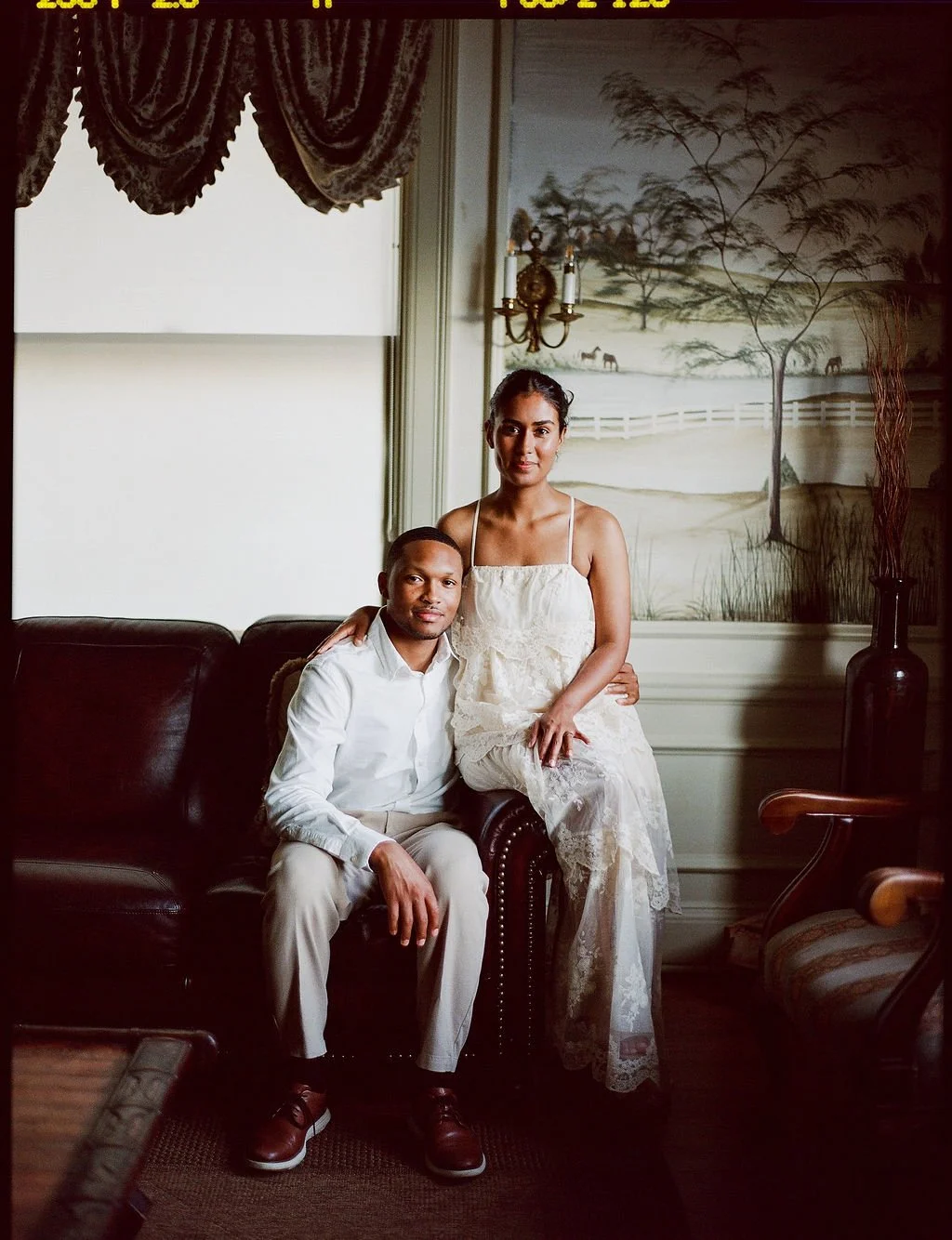 A man and woman sitting on a dark leather sofa in a vintage-style room. The man is seated, wearing a white shirt and light-colored trousers, and the woman is standing beside him with her arm around his shoulder, wearing a white lace dress. There is a landscape painting with trees, horses, and a fence on the wall behind them, along with a wall-mounted sconce and a tall vase with dried branches on the right.