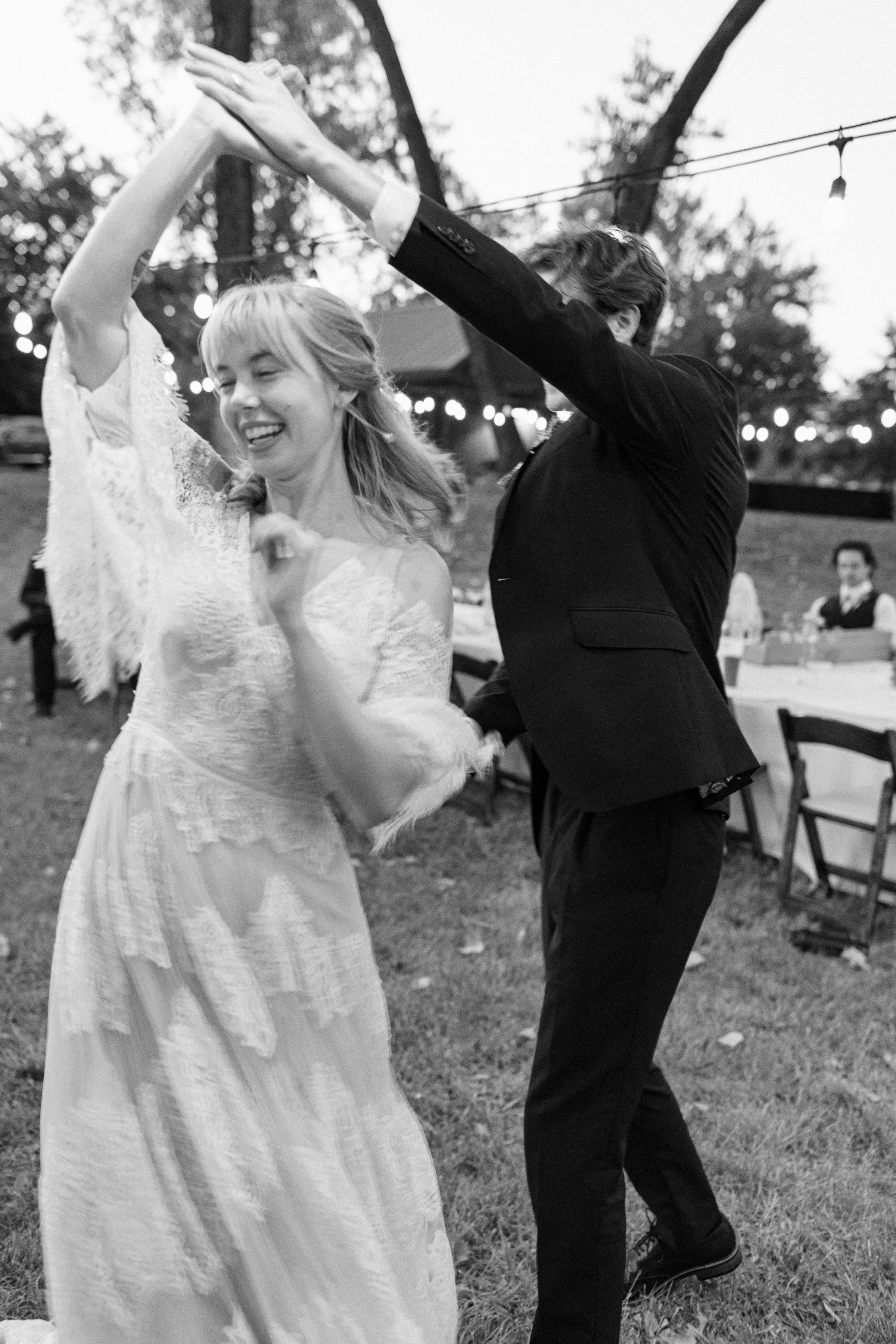Couple dancing at an outdoor wedding reception with string lights, smiling woman in a lace dress and a man in a suit.