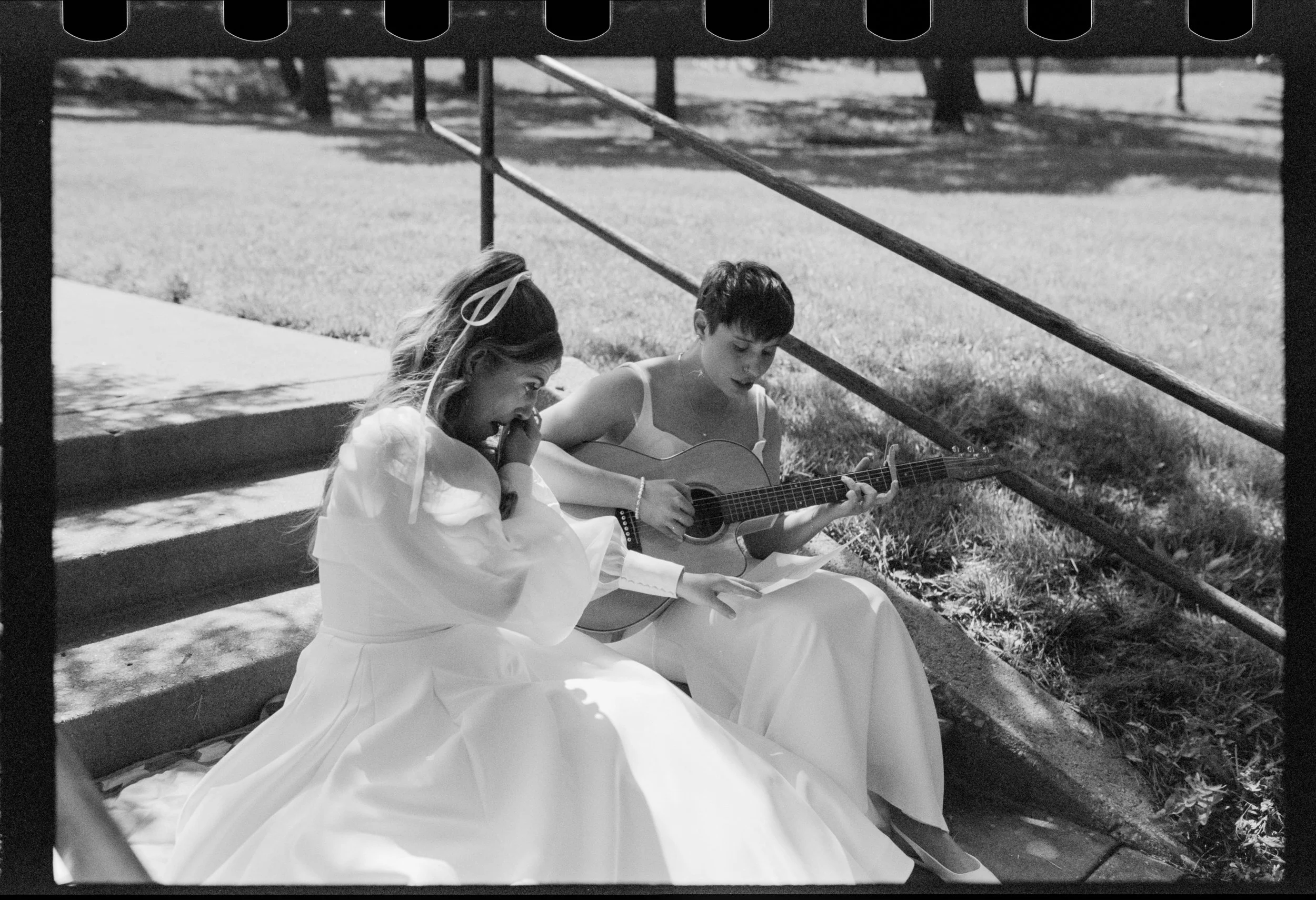 Black and white photo. One woman is listening to her partner sing to her, looking excited and crying tears of joy. The other woman is playing an acoustic guitar.