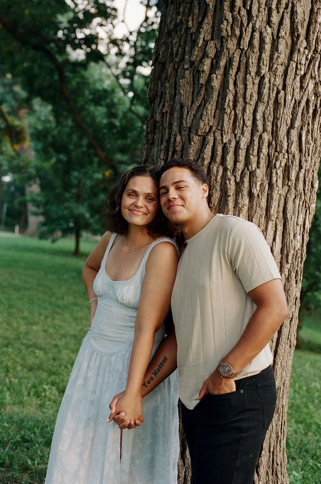 A couple standing close together outdoors next to a large tree, holding hands and smiling, with green grass and trees in the background.