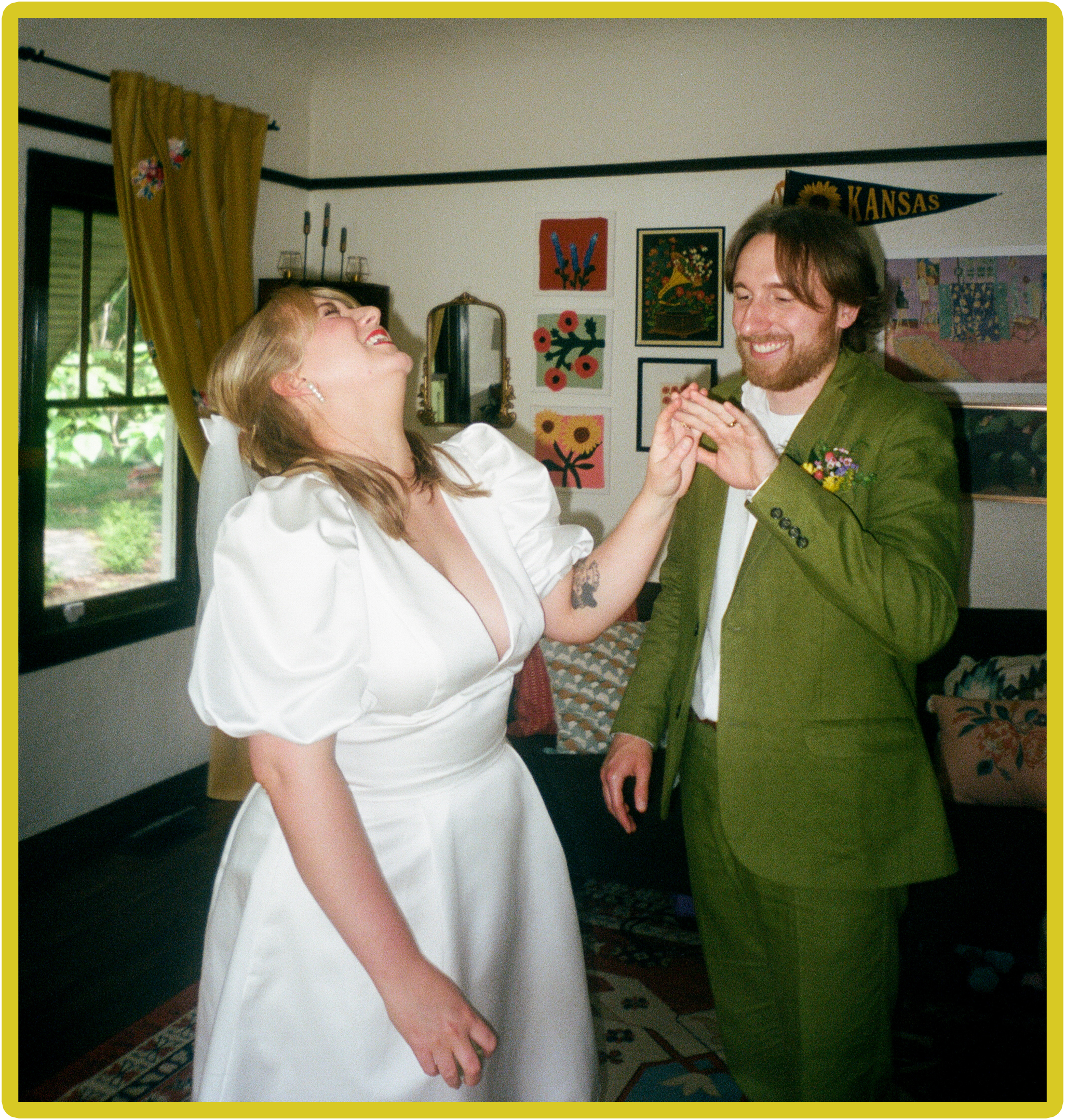 A couple celebrating a wedding, with one partner in a white bridal gown and the other in a green suit, holding hands and smiling inside a decorated room.