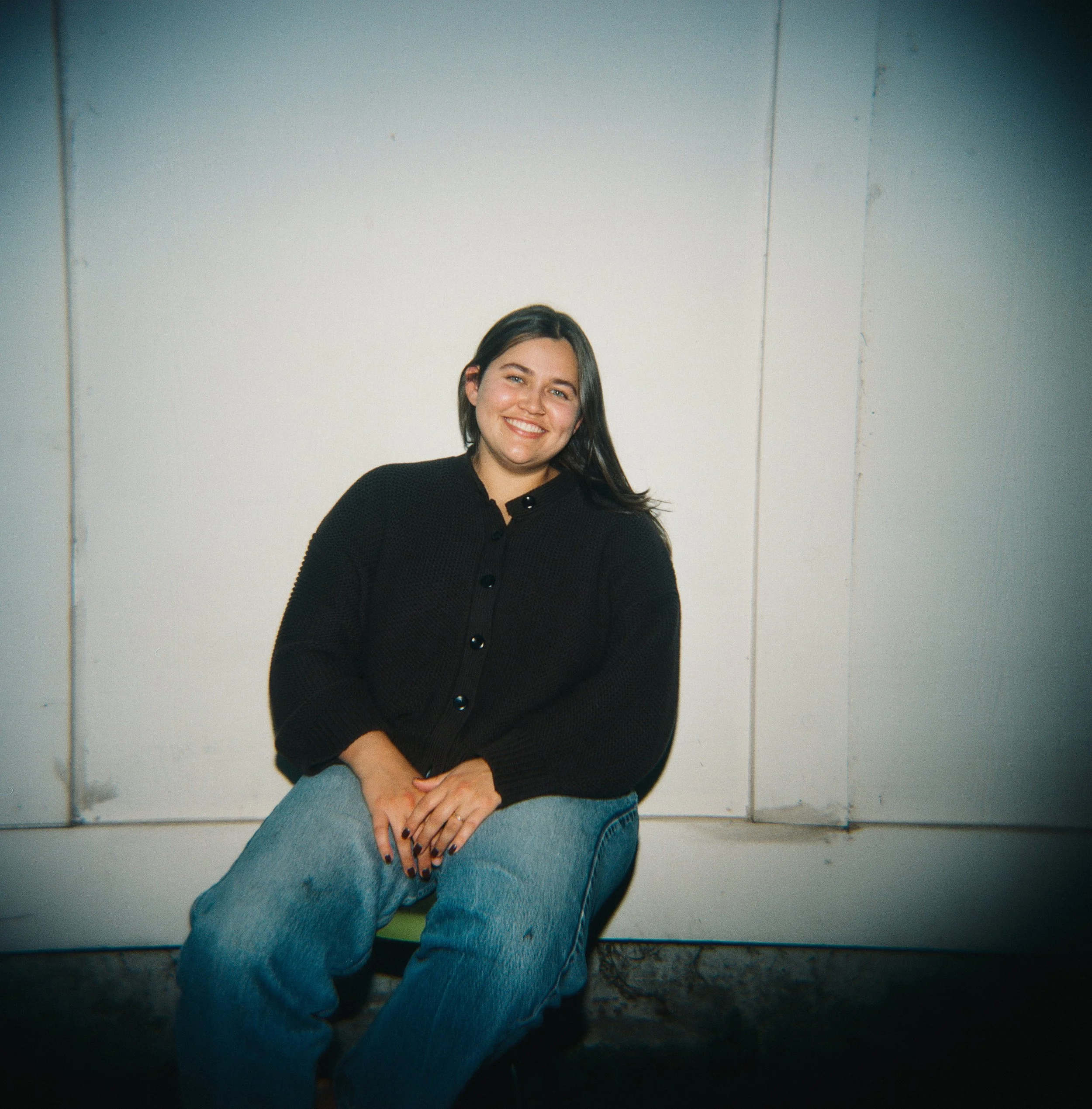 Delaney Hady, smiling, wearing a brown sweater and blue jeans, seated against a plain white wall