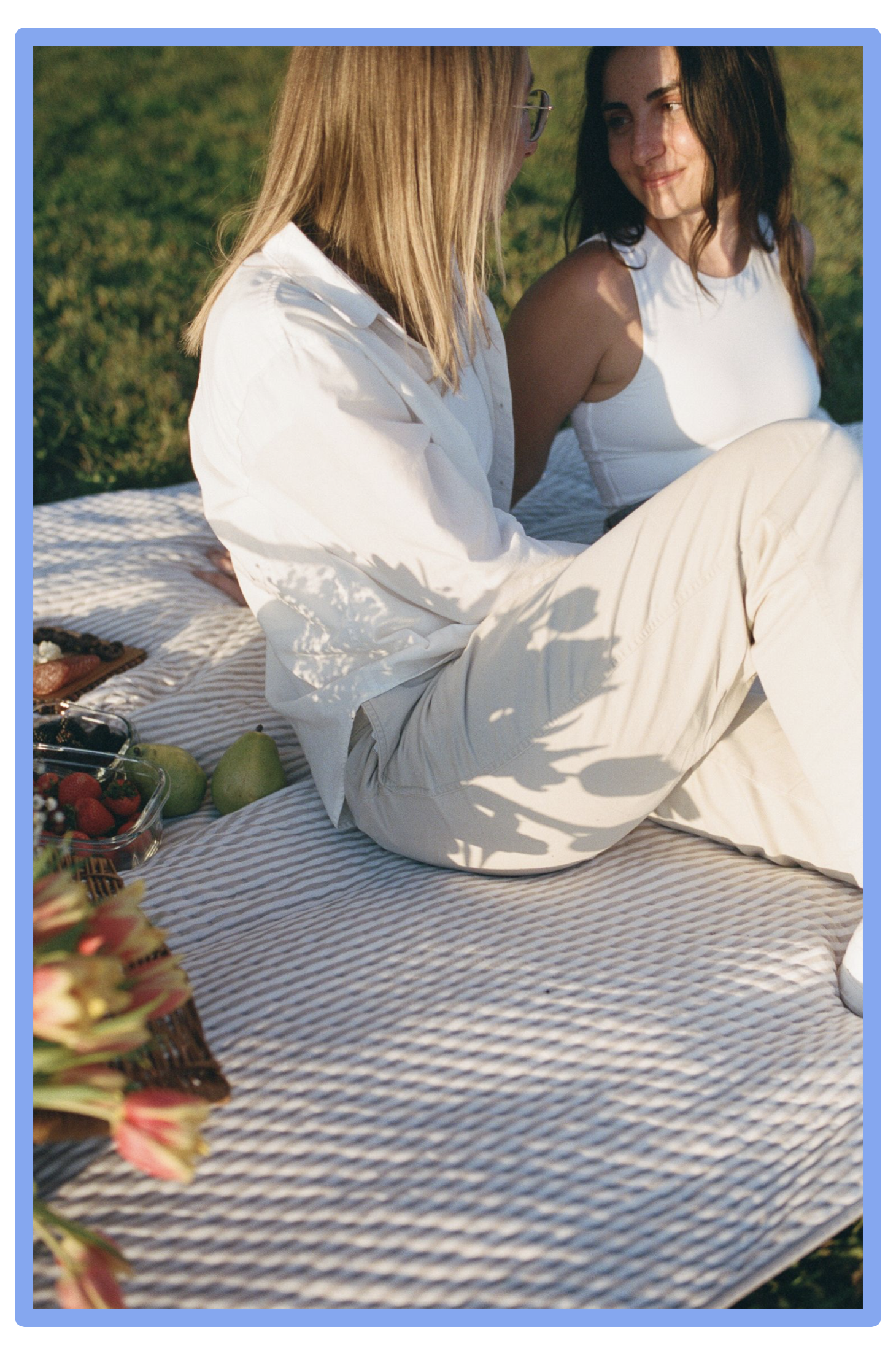A couple sitting on a picnic blanket outdoors, having a picnic with fruits and flowers, during daytime.
