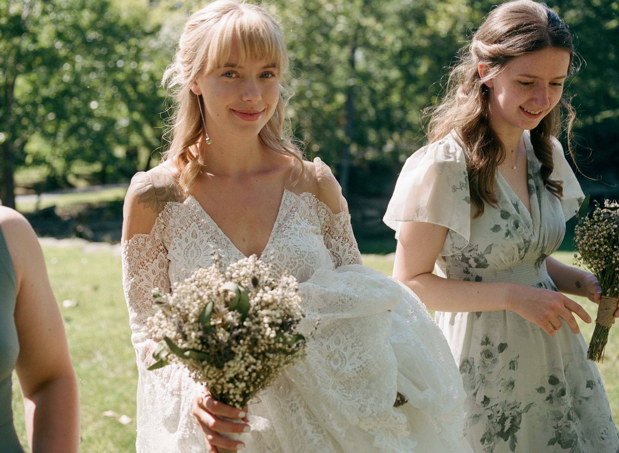 A bride and her bridesmaids holding bouquets walk outdoors in a sunny park, one in a white lace dress and the other in a floral dress.