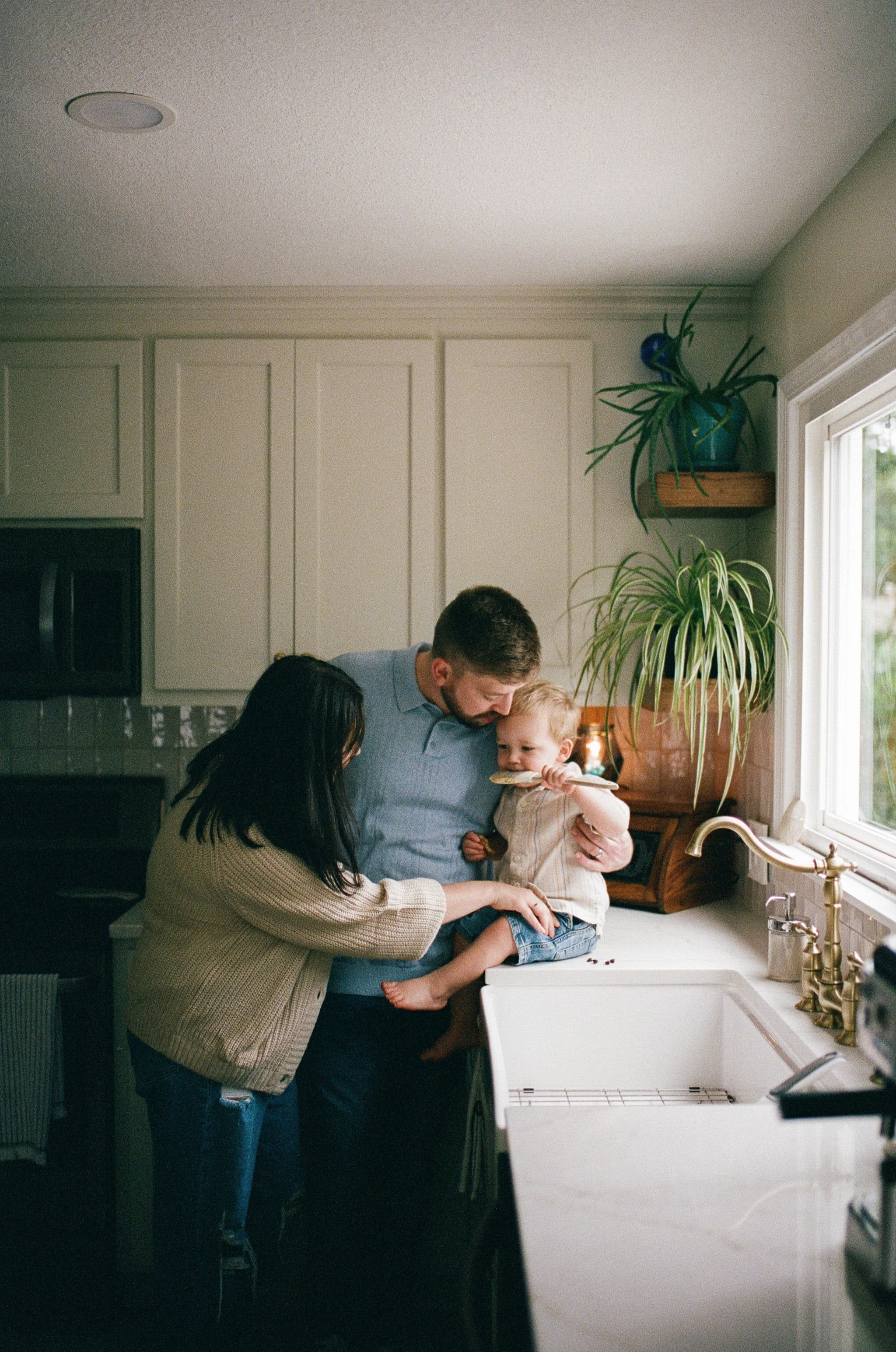 A family of three in the kitchen with a father, mother, and young son. The mother helps the son, who is sitting on the kitchen counter, eat cookies while the father holds him. The kitchen is bright with sunlight, decorated with green plants.
