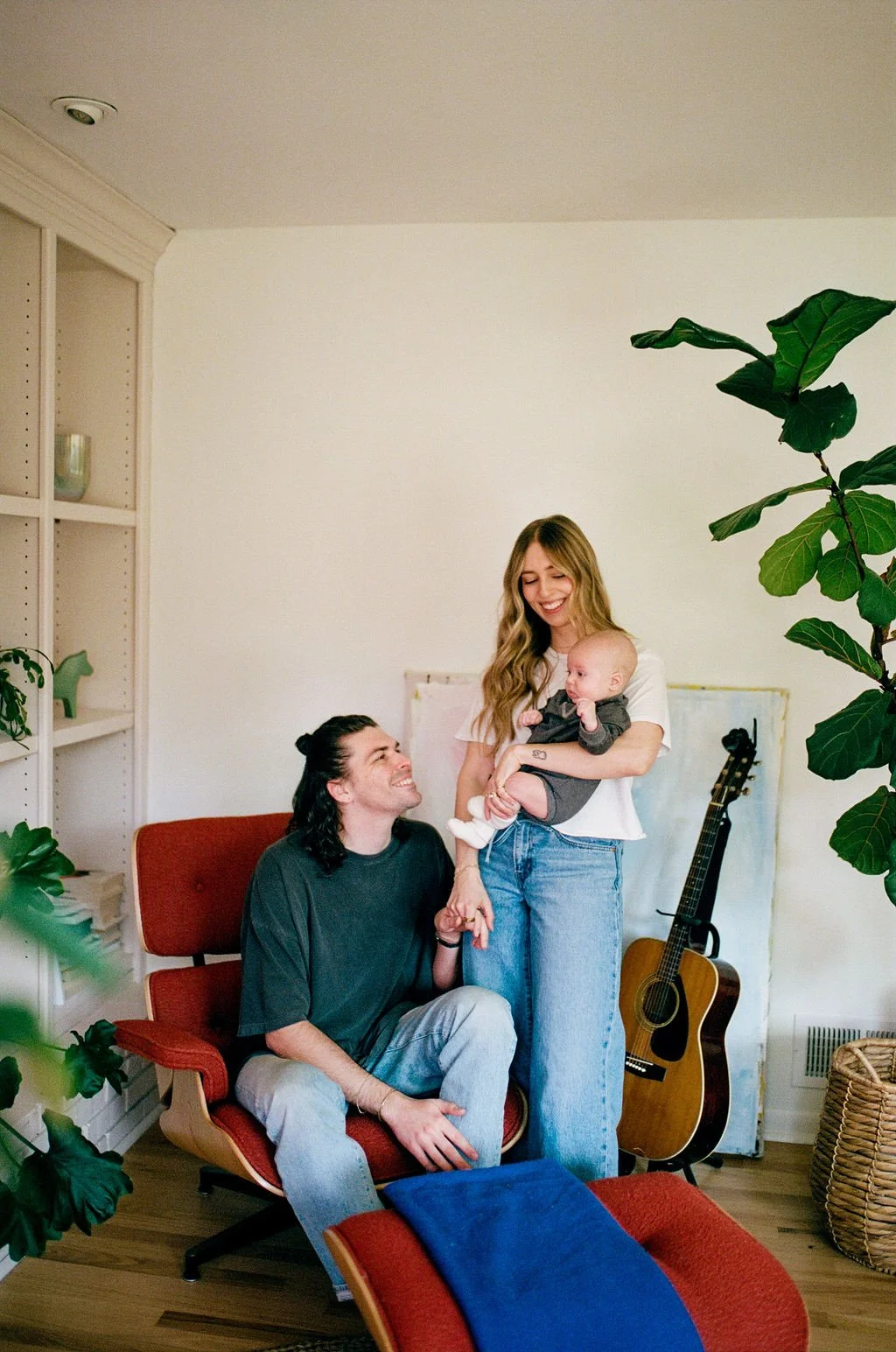 A young woman with long wavy hair holding a baby and a man sitting in a red chair in a cozy living room. The woman is smiling, and the man is looking up at her with a smile. There is a guitar leaning against the wall and a bookshelf with decorative items on the left.