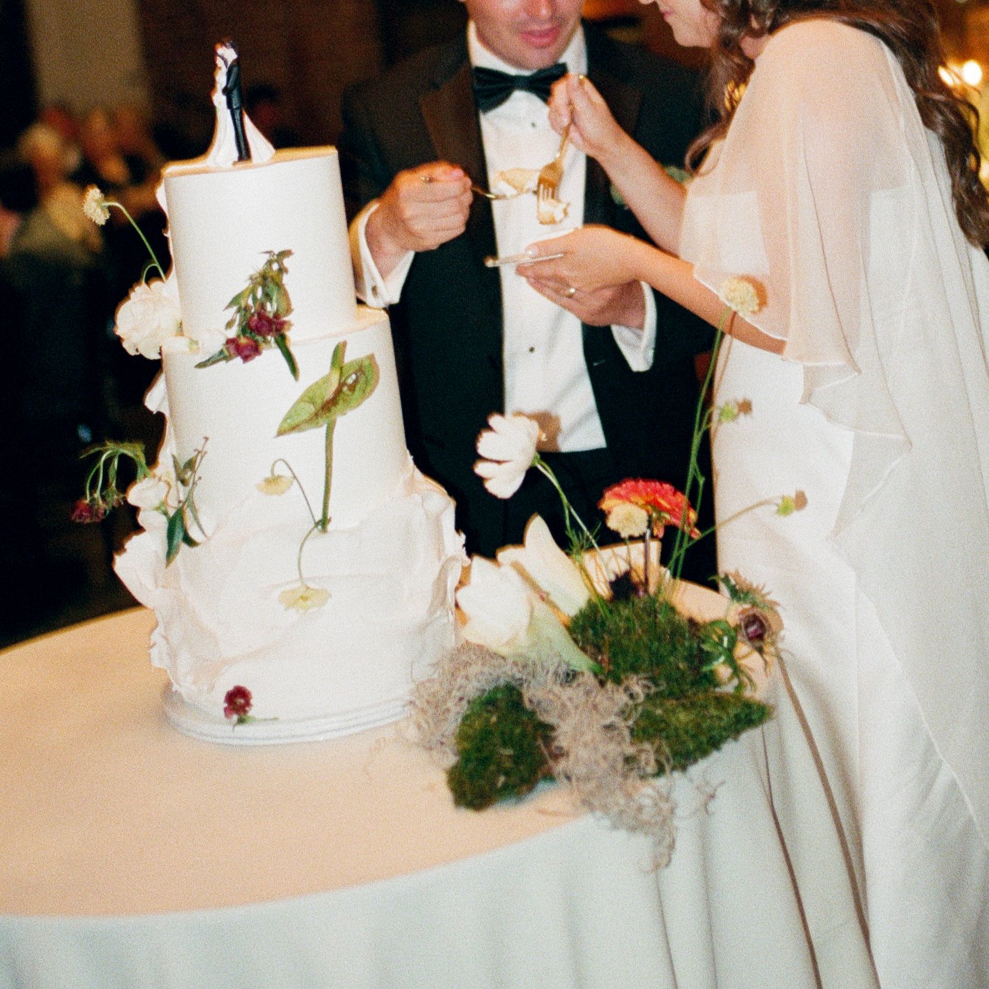 A bride and groom cutting their wedding cake together at the reception, with the cake decorated with flowers and greenery.