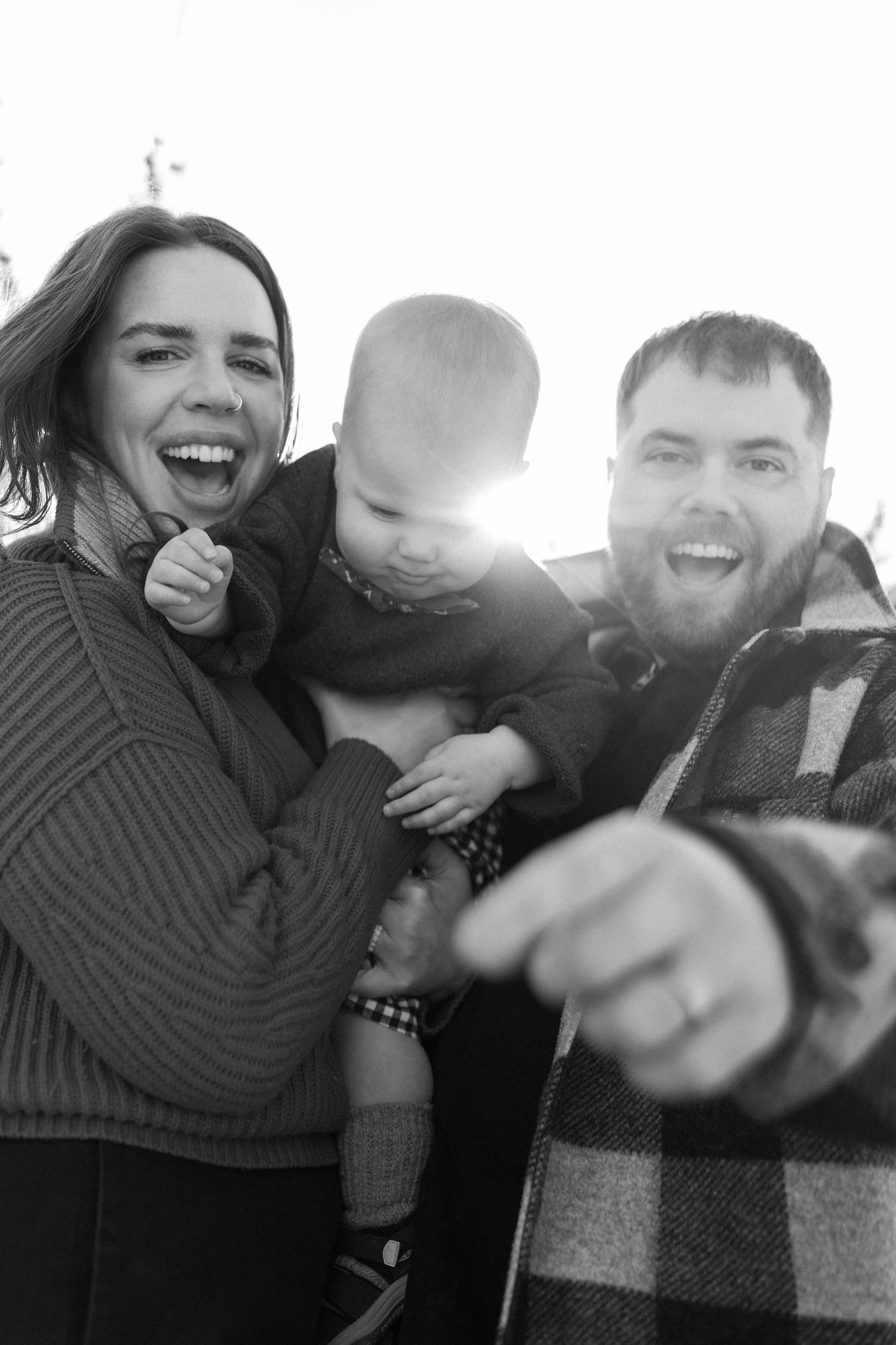 A smiling woman holding a young child, with a man pointing towards the camera, in an outdoor setting with the sun shining behind them.