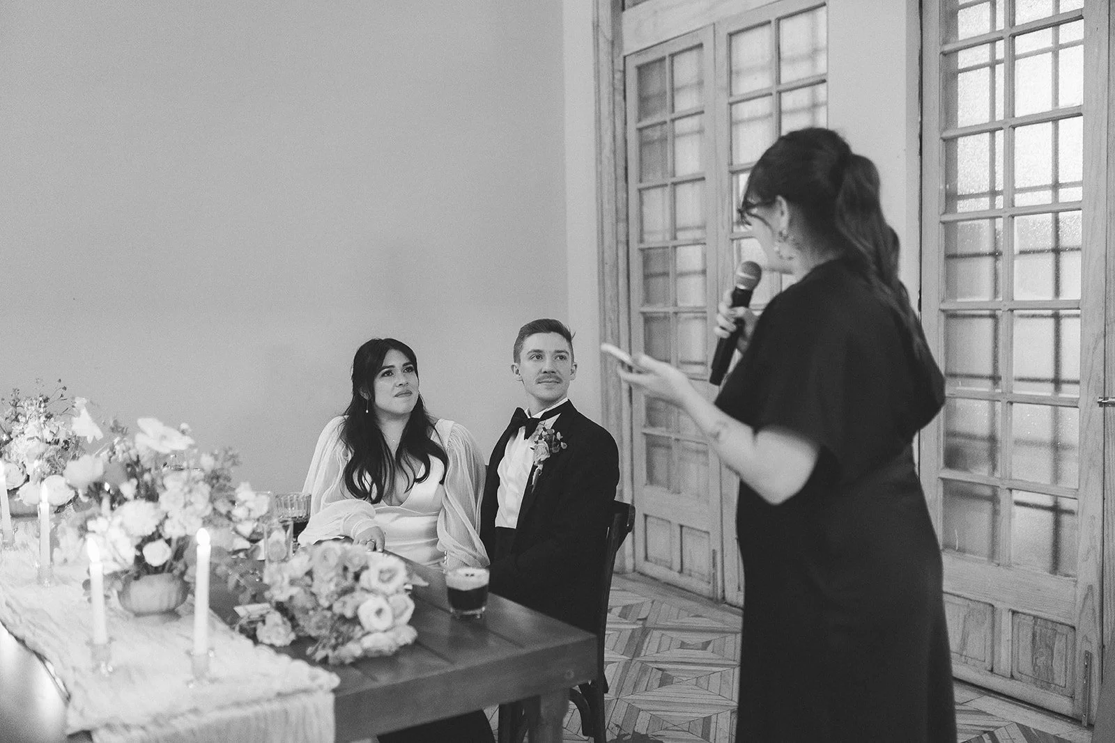 A woman gives a speech with a microphone during a wedding reception. The bride and groom are seated at a decorated table and listening attentively.