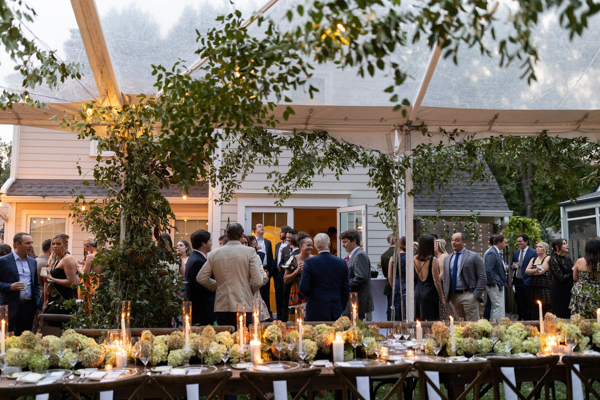 Guests socializing at an outdoor party or wedding reception under a canopy, with a long table decorated with candles and flowers in the foreground.