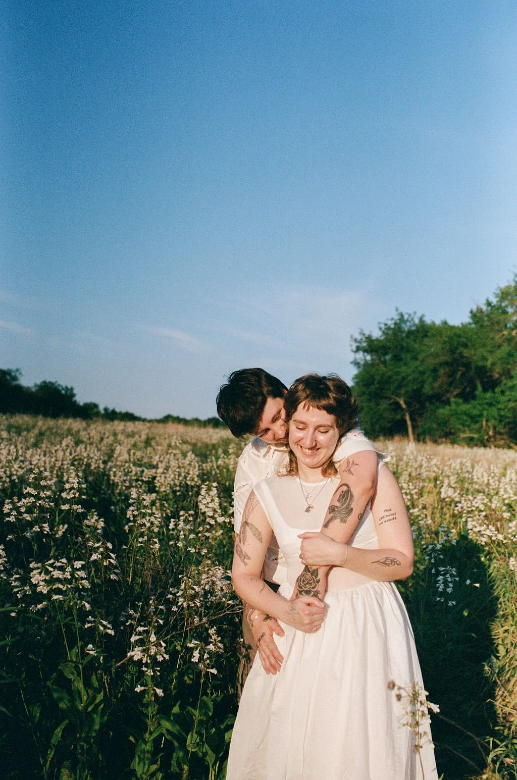 A queer couple embraces and smiles in a field of white flowers under a clear blue sky.