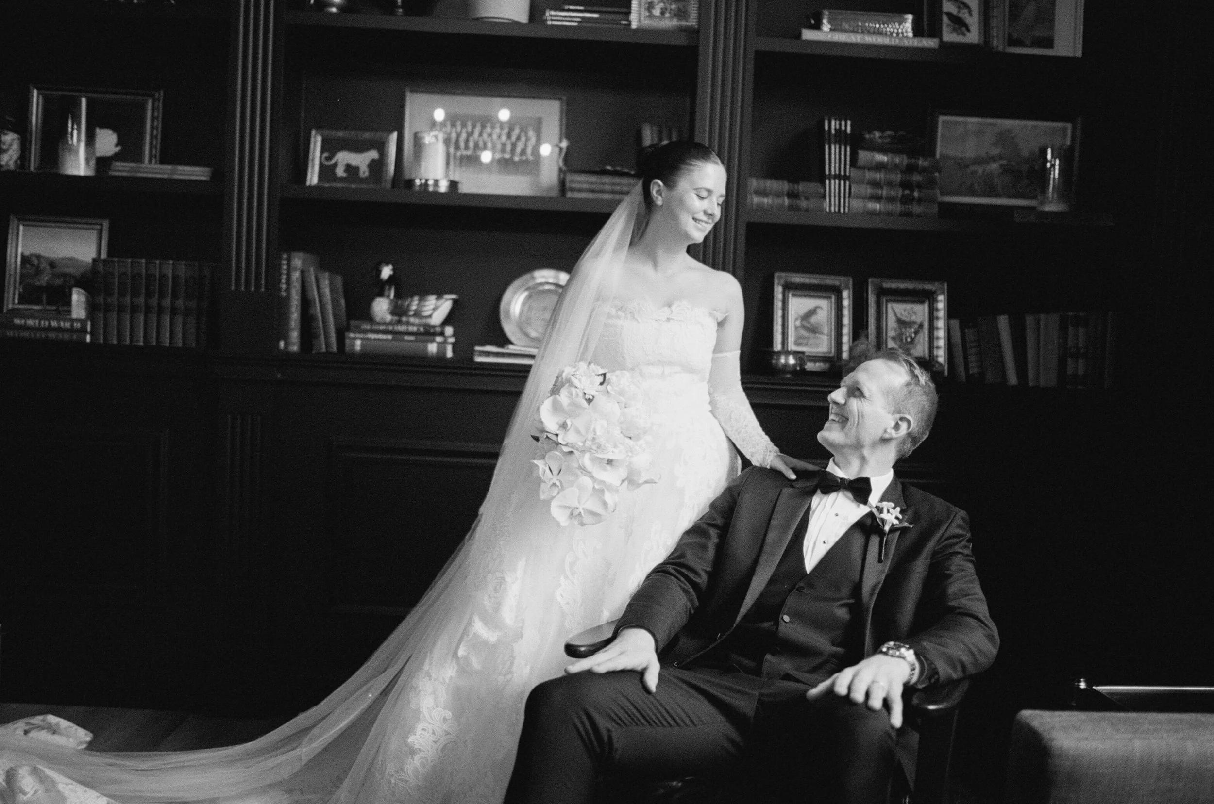 A bride in a wedding gown with a long train and veil stands smiling at her husband in a tuxedo sitting in a chair in a room with dark bookshelves and framed pictures.