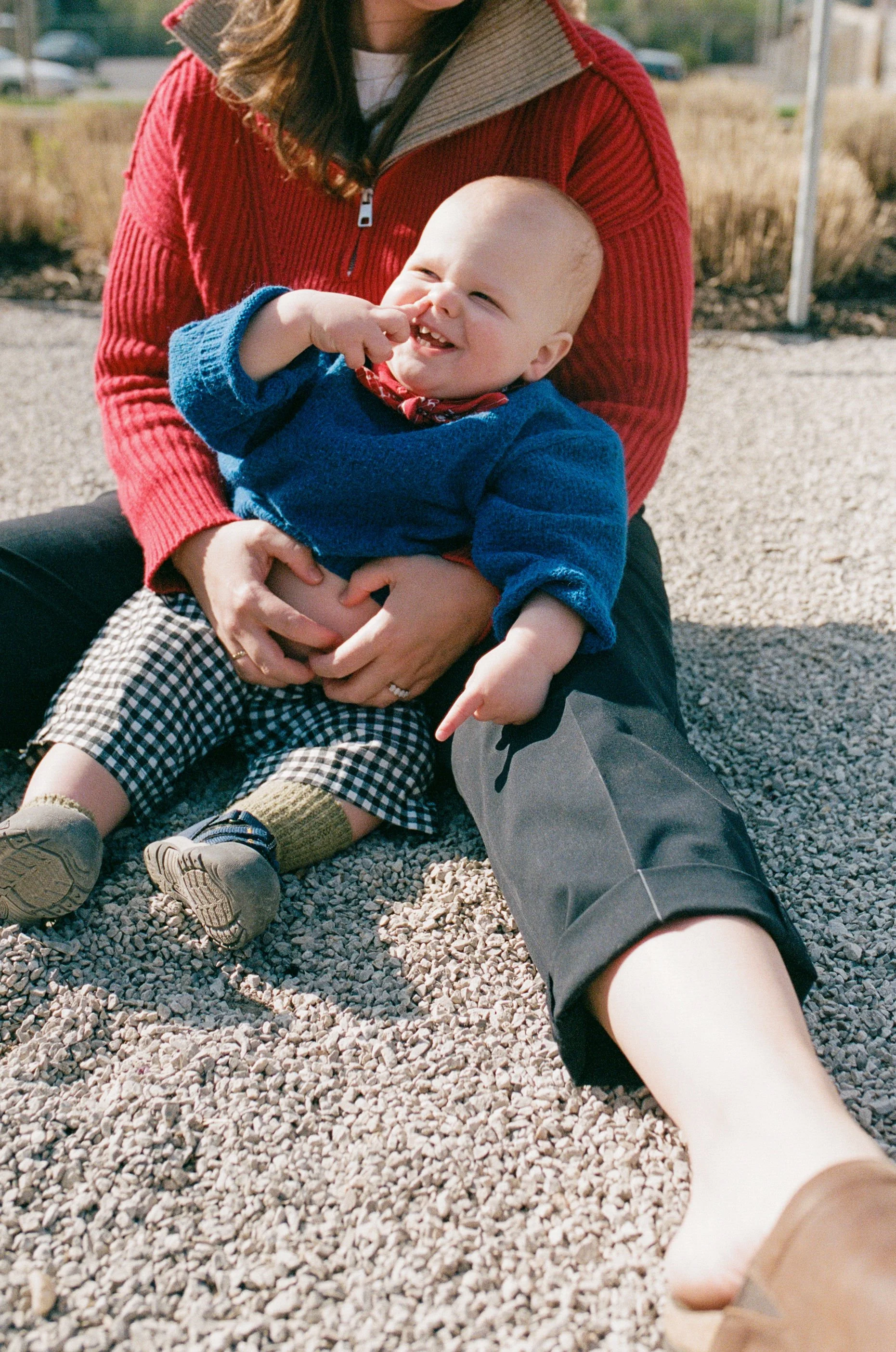 A woman and a young child sitting outdoors on a gravel surface, with the woman holding the child on her lap. The child is smiling and picking his nose.