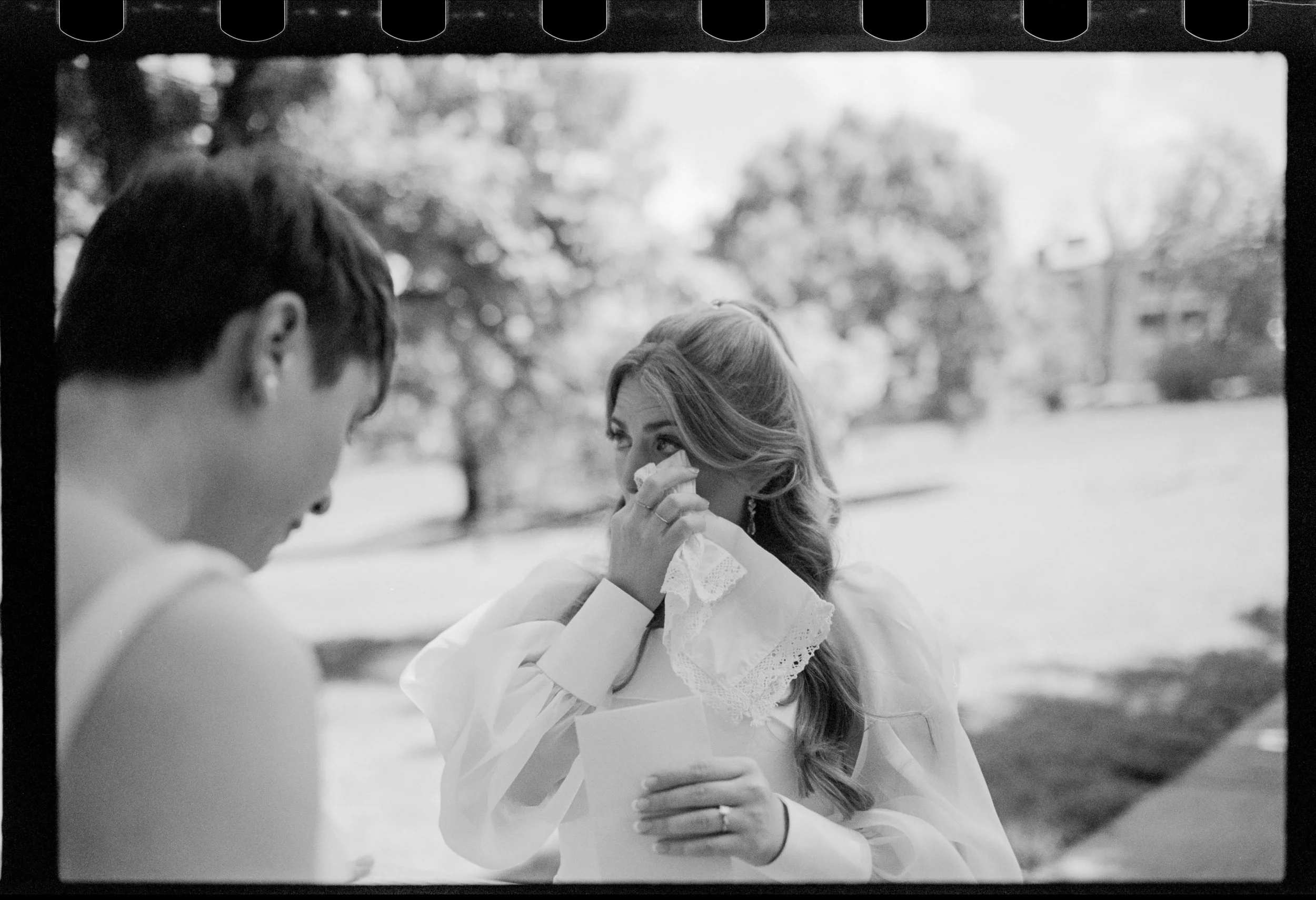 A bride in a white dress wiping a tear from her eye while her wife, also dressed in white, looks on. She is holding her vows and listening to her wife share what she's written.
