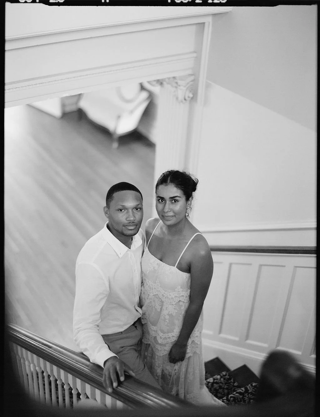 A black and white photo of a couple standing on a staircase looking up at the camera.