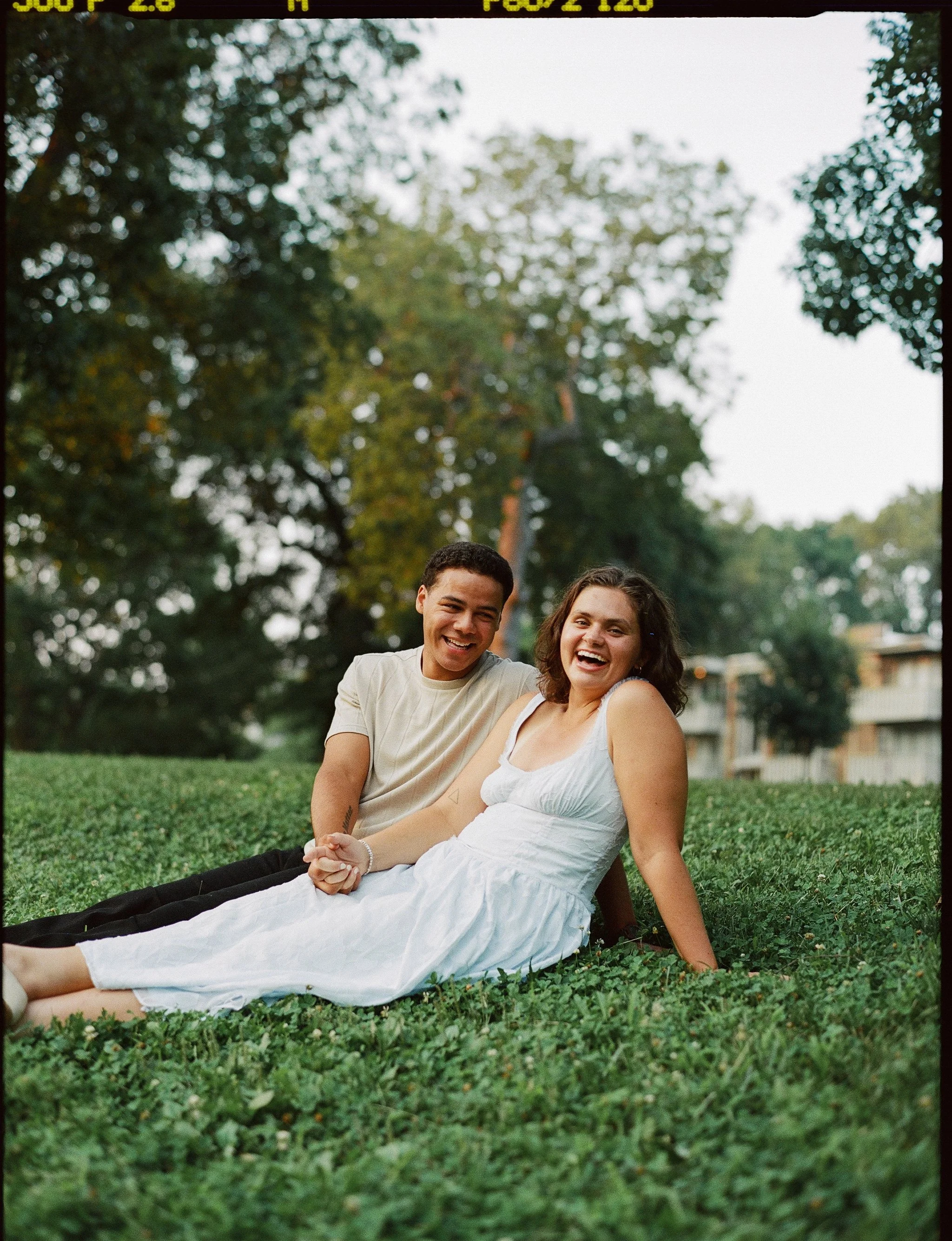 A young man and woman sitting on grass outdoors, smiling and holding hands. The woman is wearing a blue dress, and the man is wearing a beige shirt and black pants. Trees and a building are visible in the background.