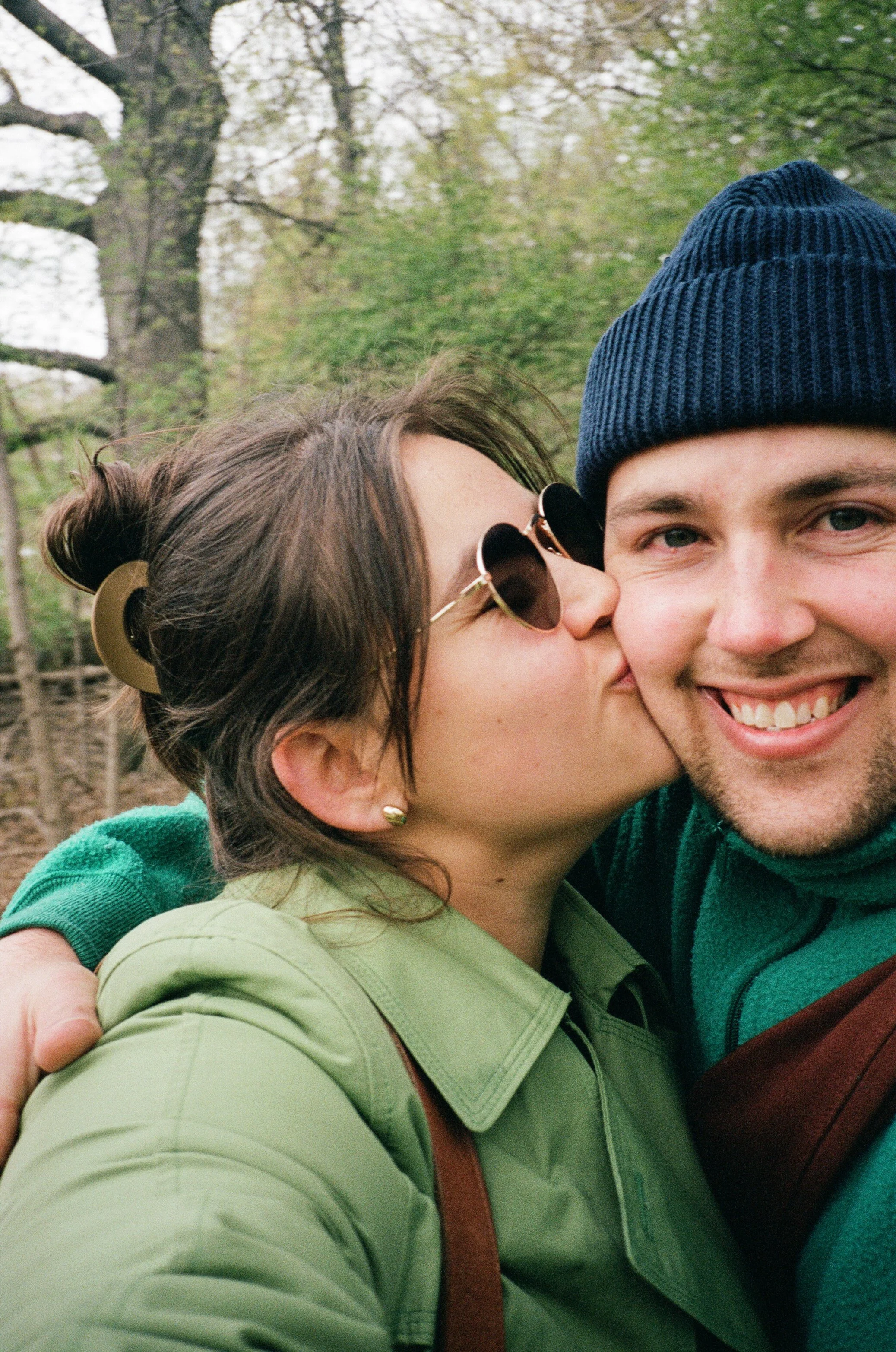 A woman kisses a man on the cheek in a park with trees in the background.