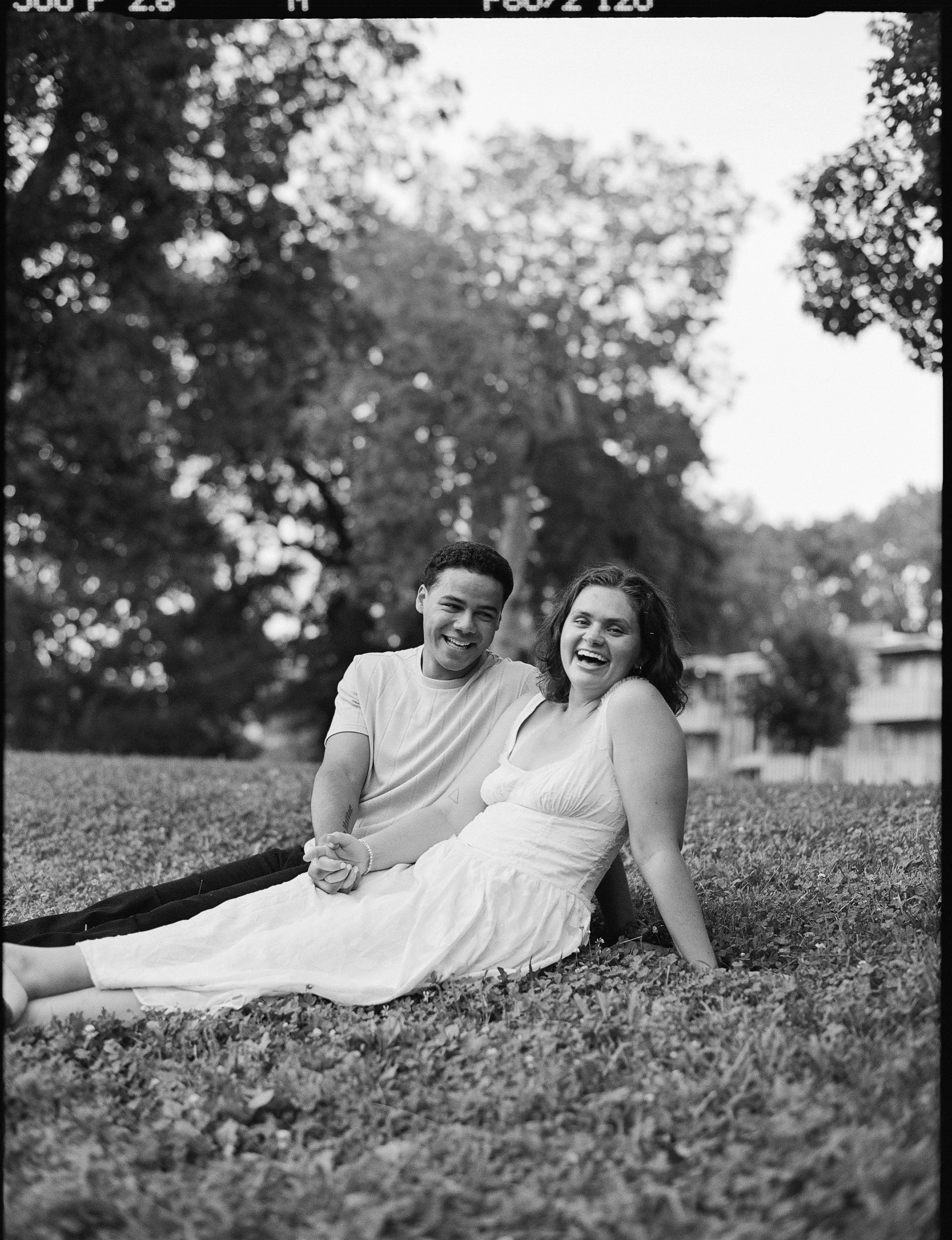 A black-and-white film photo of a young man and woman sitting on the grass outdoors, holding hands, and smiling.