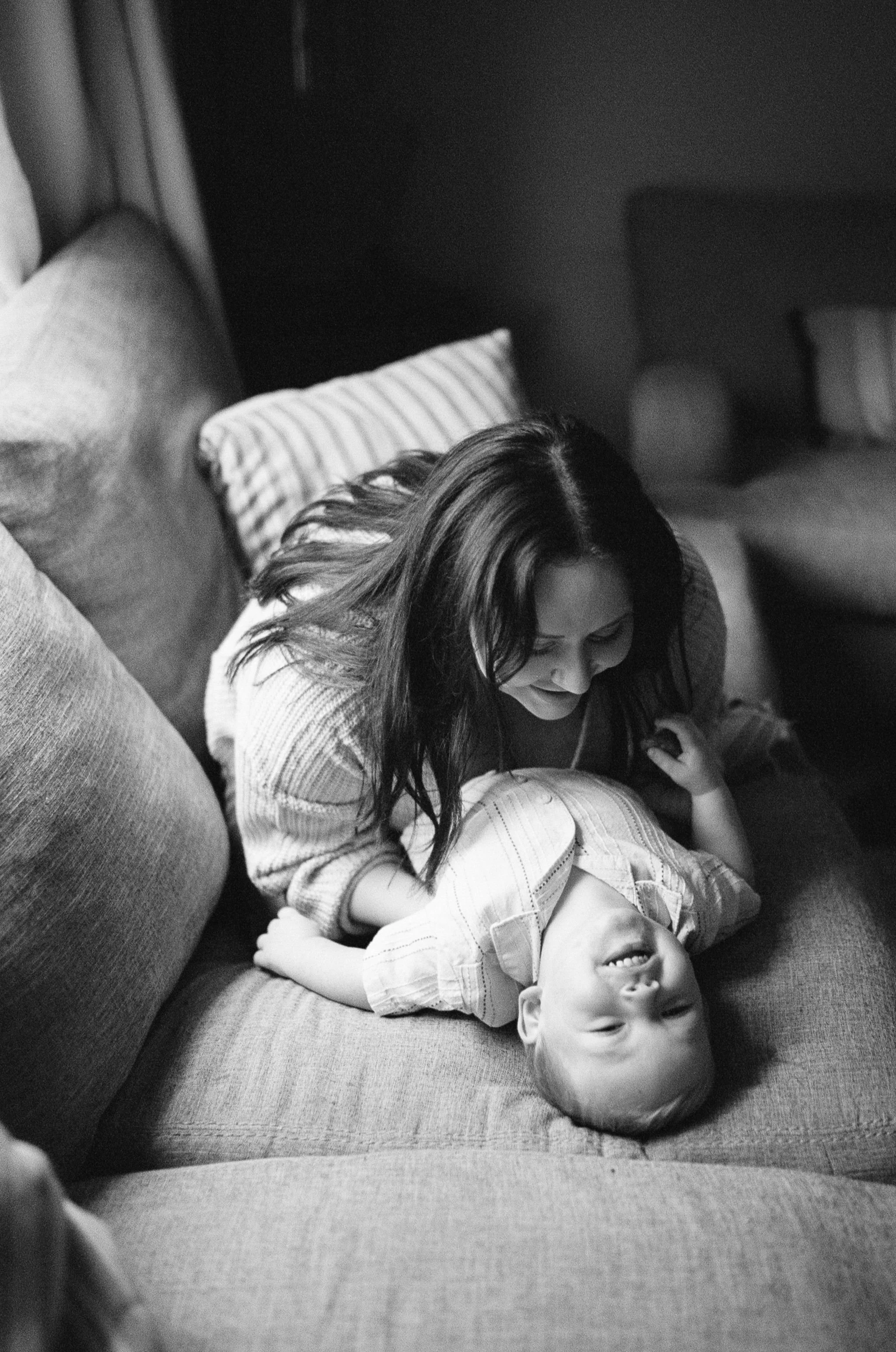 A woman playfully wrestling with her son on a couch. The boy is lying on his back, smiling, while the woman leans over him, both appearing to enjoy a playful moment.