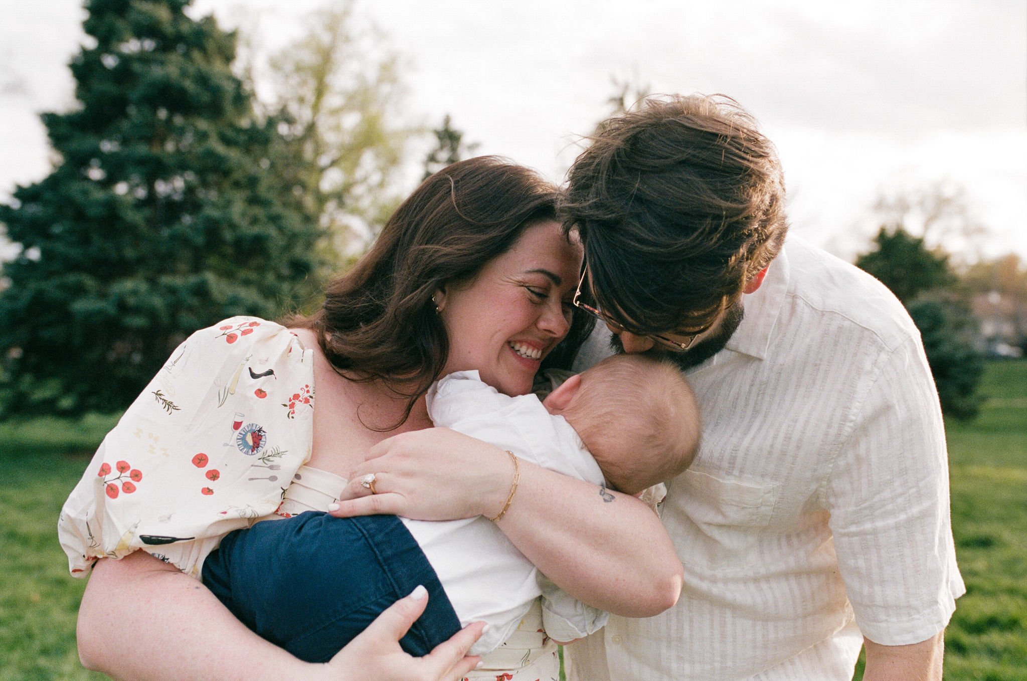A happy family of three, a woman holding a baby, a man leaning in, outside in a park with trees in the background.