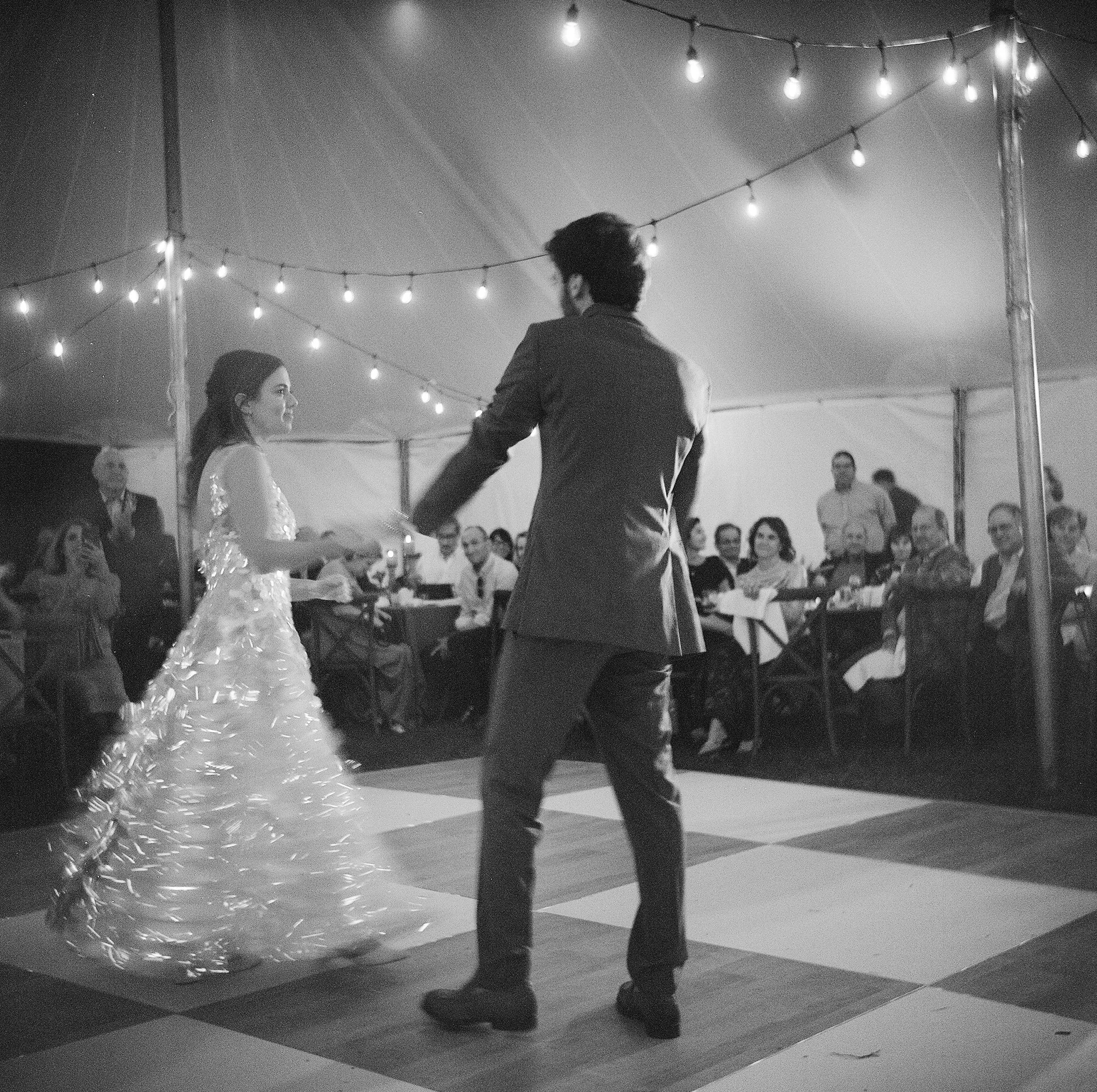 A bride and groom dancing at their wedding reception under a tent with string lights, surrounded by seated guests.